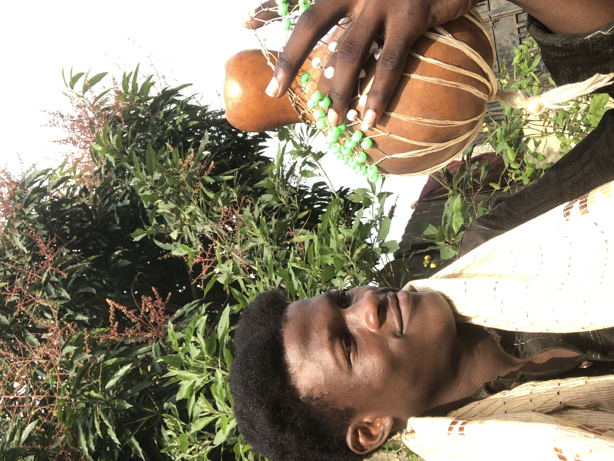 A young Black man with dark skin and short, curly black hair is pictured outdoors, looking to his left with a slight smile. He is wearing a black shirt and a patterned white and gold scarf around his neck. His right hand, with manicured fingernails, is holding a dried gourd instrument adorned with green and white beads strung on twine. The gourd is tied with multiple strands of twine, and a braided white tassel hangs from its bottom.
The man is positioned in the foreground, with lush green foliage and branches from a tree, possibly a mango tree with developing fruit clusters, forming the background. The lighting suggests it is daytime, with the sky appearing bright and washed out. The overall scene suggests a natural outdoor setting, potentially in a garden or yard in Ado, Nigeria.