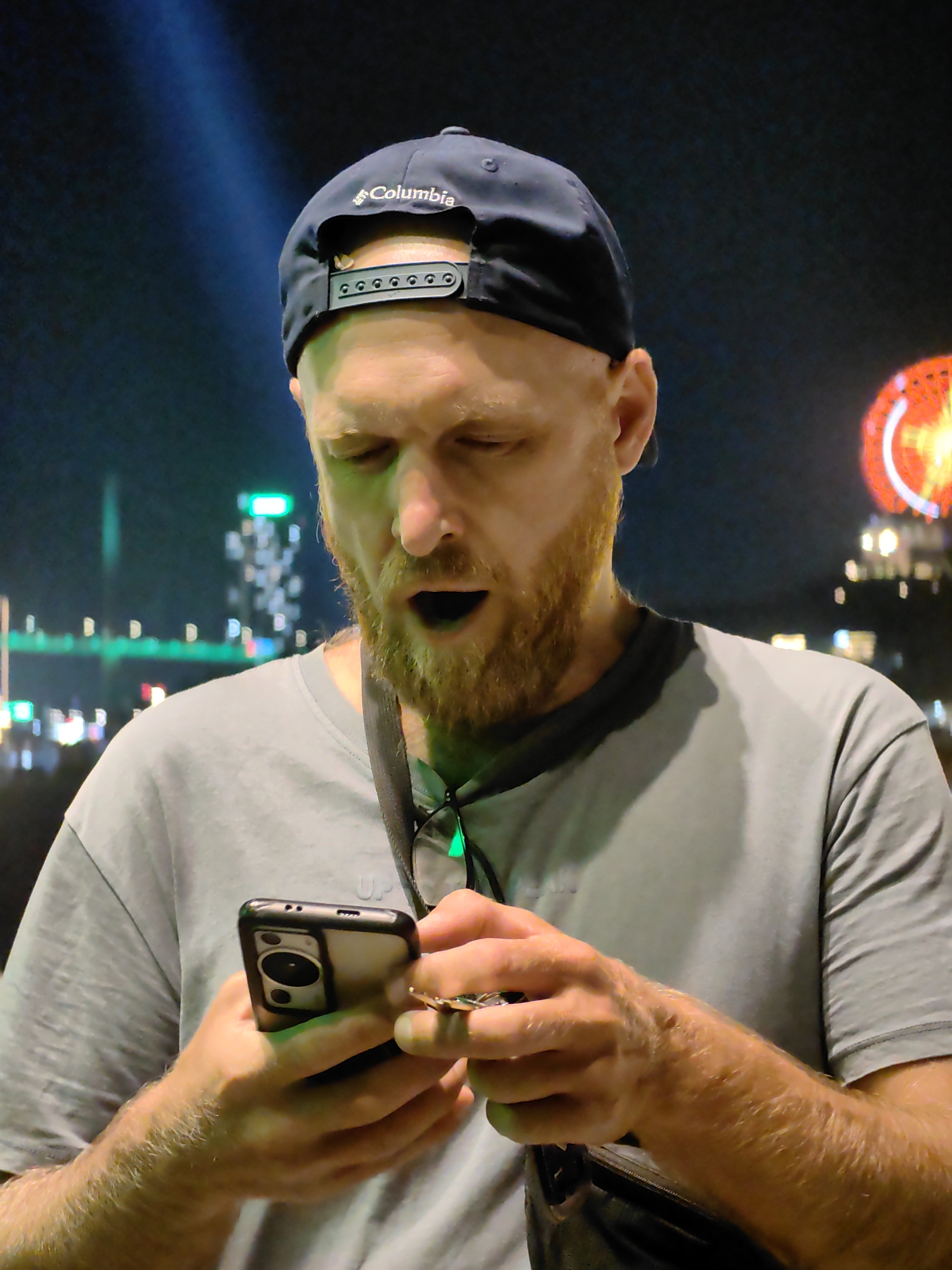 A medium shot depicts a Caucasian male with red hair and a beard, wearing a blue Columbia baseball cap, a gray t-shirt, and a black bag. The man is looking down at a black smartphone he holds in his hands, with his mouth open in an expression of surprise. In the background, there are city lights suggesting an urban environment. Based on the presence of a Ferris wheel and illuminated buildings, the location is consistent with Bai Chay Ward, Vietnam.