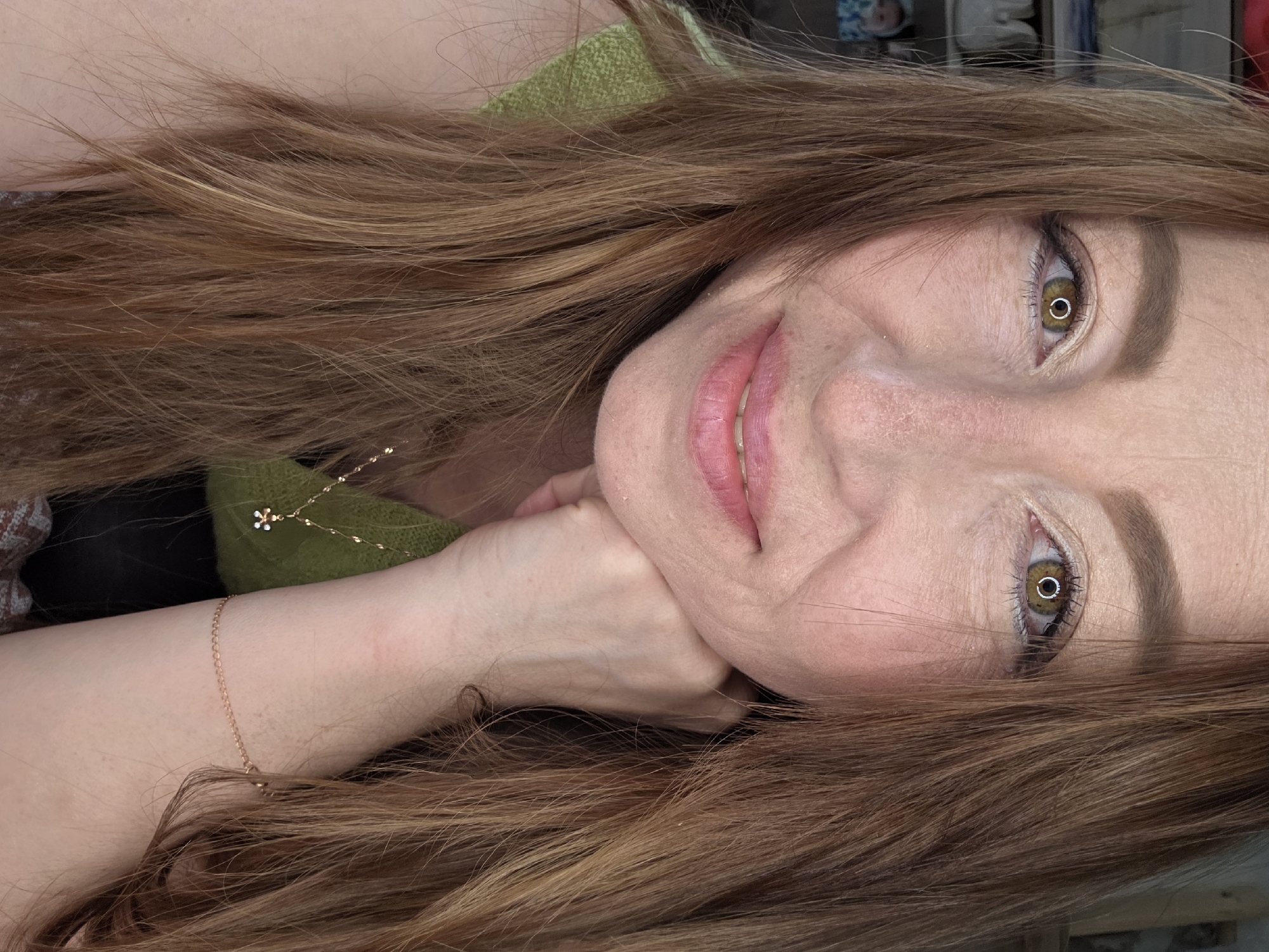 The image depicts a close-up, chest-up portrait of an adult female in an indoor setting in Chaikovsky, Russia. The individual has fair skin with visible freckles on her nose and cheeks. Her long, reddish-brown hair with lighter highlights frames her face and flows over her shoulders. She possesses light hazel or brown eyes, which show prominent circular reflections from a direct artificial light source. Her eyebrows are well-groomed and light brown, and her eyelids are adorned with light-colored eyeshadow. Her lips are a natural pink hue, slightly parted to reveal her upper teeth, forming a subtle smile. Her right hand is visible, clenched lightly, with knuckles resting beneath her chin. She wears a lime green, possibly knitted, sleeveless top. A delicate gold-colored chain necklace with a small, intricate charm (appearing to be a light-colored figure with white and blue accents) is visible on her left side, and a thin gold-colored chain bracelet is on her right wrist. The background is dark and heavily out of focus, showing indistinct shapes on the left side, possibly shelving with light-colored objects and a blurred image of a child. The woman is looking directly at the viewer.