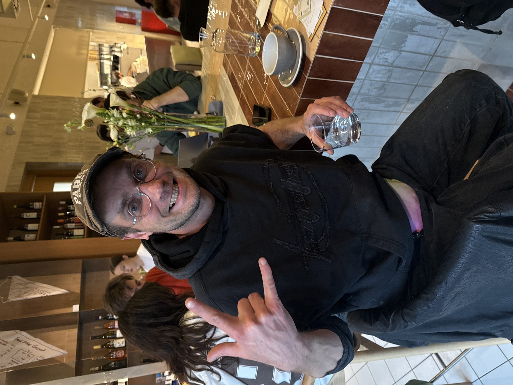 The image depicts a male individual seated at a tiled table within a cafe or restaurant in Belgrade, Serbia. The man, a Caucasian adult, wears a dark hooded sweatshirt with distressed embroidery, dark trousers, circular wire-frame glasses, and a dark baseball cap with "BALENCIAGA" partially visible. He is smiling broadly, making direct eye contact with the camera, and holds a clear glass of water in his left hand. His right hand is raised, forming a "rock on" gesture with the index finger and pinky extended. A dark, possibly leather, jacket is draped over the back of his wooden chair. A bright pink or purple garment is visible peeking from his waistband.
The table features brown and light-colored square tiles. On the table, to the man's right, are an empty grey ceramic coffee cup and saucer, a clear glass carafe, a dark smartphone, and a small stack of papers including one showing "2000". A glass vase containing white calla lilies and green foliage is positioned further down the table.
In the background, other patrons are visible, out of focus. To the man's left, a woman with long dark hair is seen from behind, wearing a white top. Further back, a person in a dark green hooded top is looking at a silver laptop. The establishment's interior features light-colored walls, recessed ceiling lights, and wooden shelving stocked with various bottles, likely wine or spirits. Two decorative, cone-shaped paper items are suspended from the ceiling. A kitchen area with stainless steel surfaces and a person in a white uniform is visible in the far background. The floor near the man's feet is tiled white.