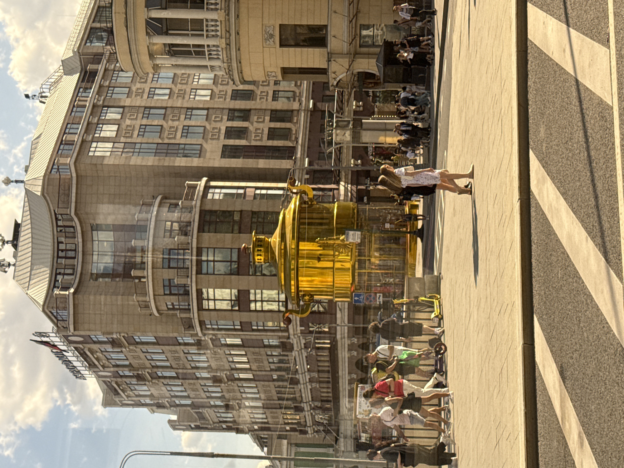 In Moscow, Russia, a bright, oversized golden samovar sculpture stands prominently in a public square. The samovar is a traditional Russian tea urn, and this replica is highly polished and reflects the surrounding environment. Behind the samovar, a large, multi-story building with an art deco architectural style dominates the background. The building is light brown with many windows and decorative elements. To the right of the samovar, another building with columns and a curved facade is visible, painted in a pale yellow color.
The foreground of the image shows a pedestrian area paved with light-colored tiles, with a crosswalk marked with white stripes on gray asphalt in front of it. Several people are walking in the square, some in casual attire, and a few are riding electric scooters. The scene is illuminated by bright sunlight, casting shadows on the ground and creating a lively atmosphere. The sky above is partly cloudy with some bright white clouds against a blue sky. The reflection on the glass in front of the observer suggests this photo was taken from inside a vehicle or building.