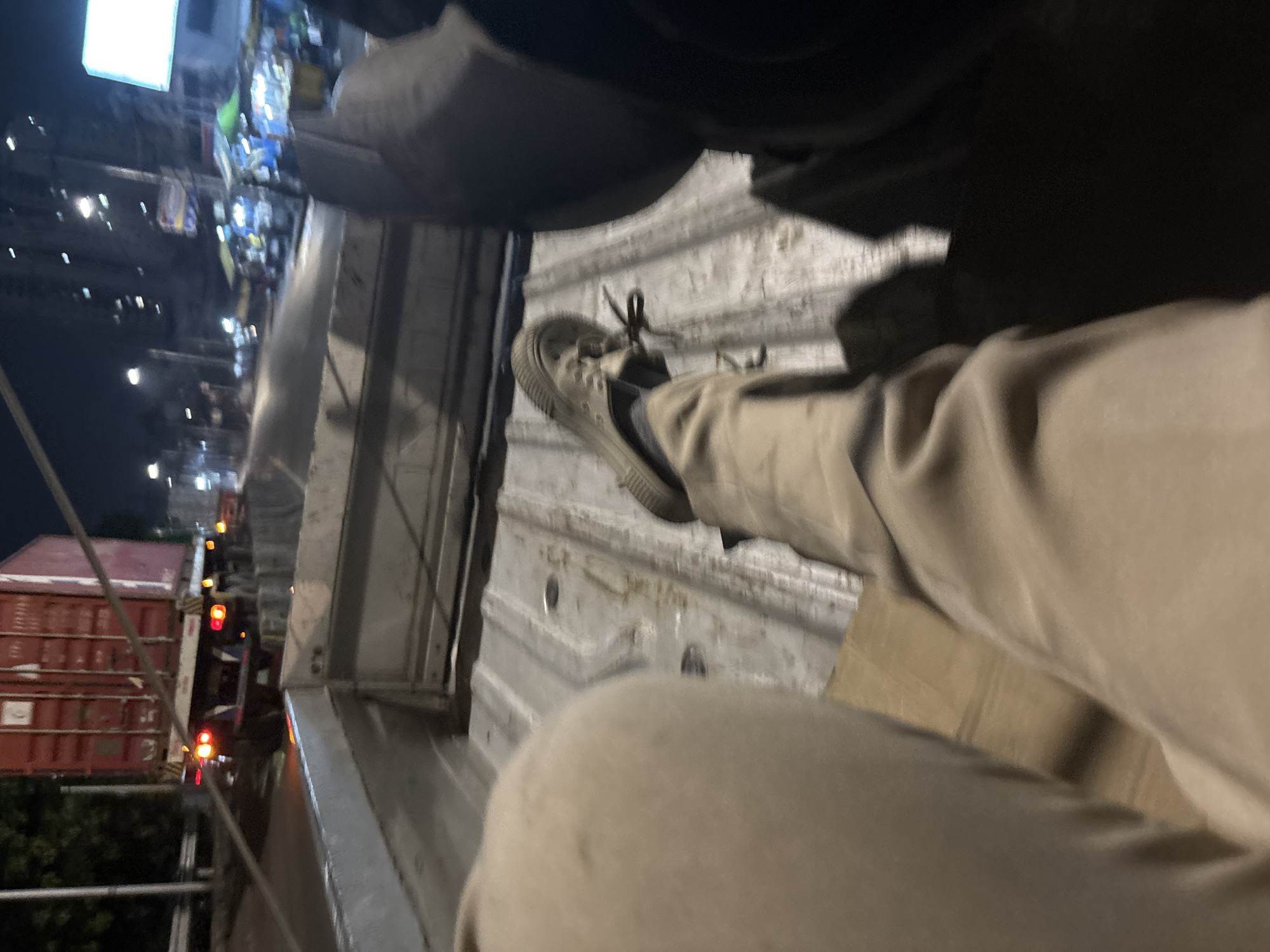 The image shows a person sitting in the back of a vehicle, possibly a truck or pickup. The person is wearing tan pants and a gray shoe. The inside of the vehicle bed appears to be metal with visible wear and tear. In the background, an urban environment at night is visible, with lights from buildings and vehicles. A red shipping container and other vehicles with red lights can also be seen. The location is identified as North Jakarta, Indonesia based on the presence of shipping containers and general architecture.