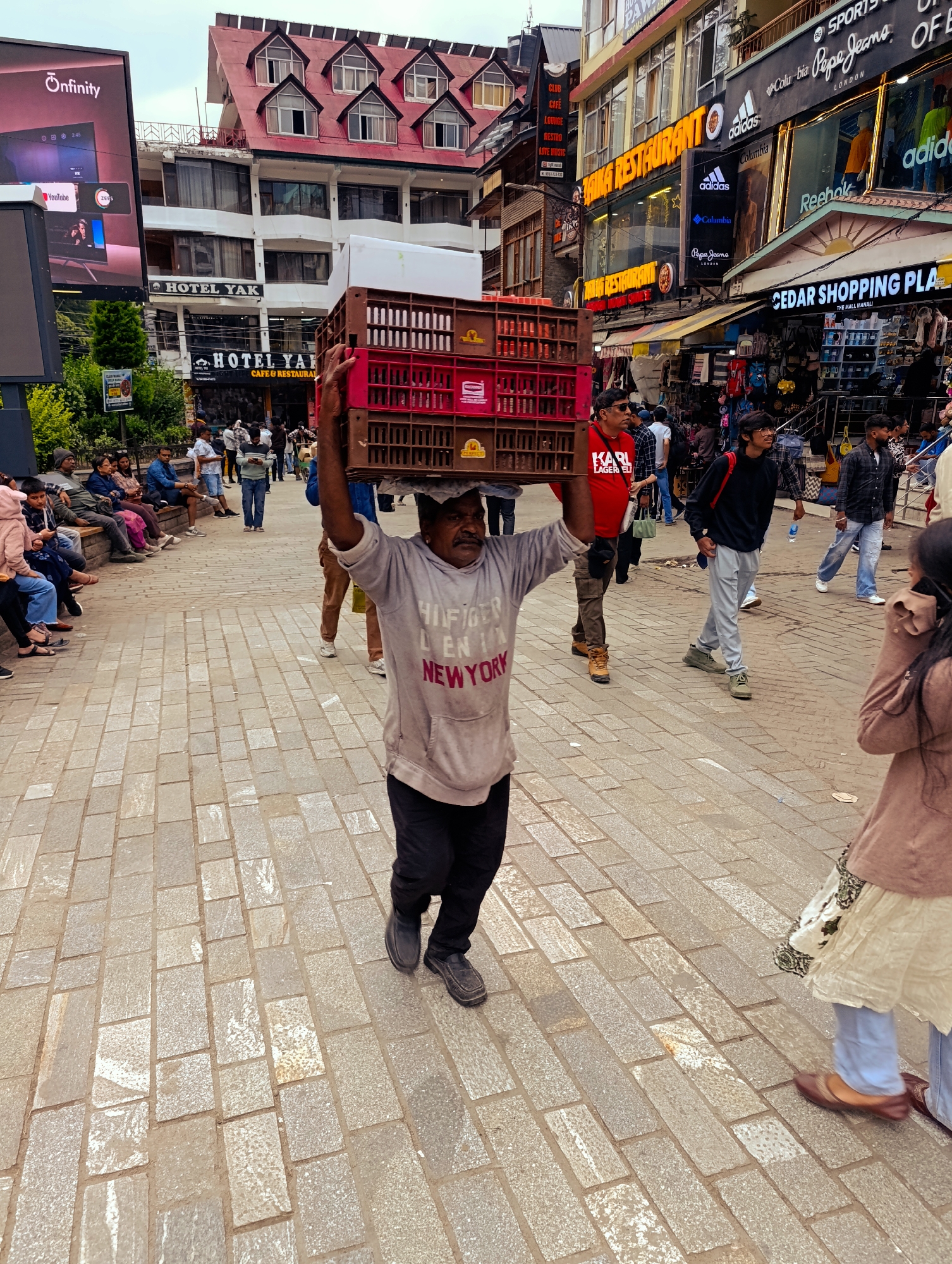 The image depicts a bustling street scene in Manali, India, likely on The Mall Road, a popular pedestrian thoroughfare. In the foreground, a man is walking with two stacks of plastic crates balanced on his head, his arms extended to maintain stability. He is wearing a grey hooded sweatshirt with text that reads "HILFIGER DENIM NEW YORK," black pants, and dark shoes. The street is paved with interlocking grey stone blocks, and to the left, a large digital billboard displays various logos and content.
In the background, various shops and buildings line the street, adorned with signage for businesses such as "HOTEL YAK," "HOTEL YAK CAFE & RESTAURANT," and "CEDAR SHOPPING PLAZA." Several people are visible walking, sitting, and interacting, creating a lively atmosphere. The architecture of the buildings, with their red-tiled roofs and multiple windows, is characteristic of the region. The overall scene conveys a sense of daily life and commerce in this tourist destination.