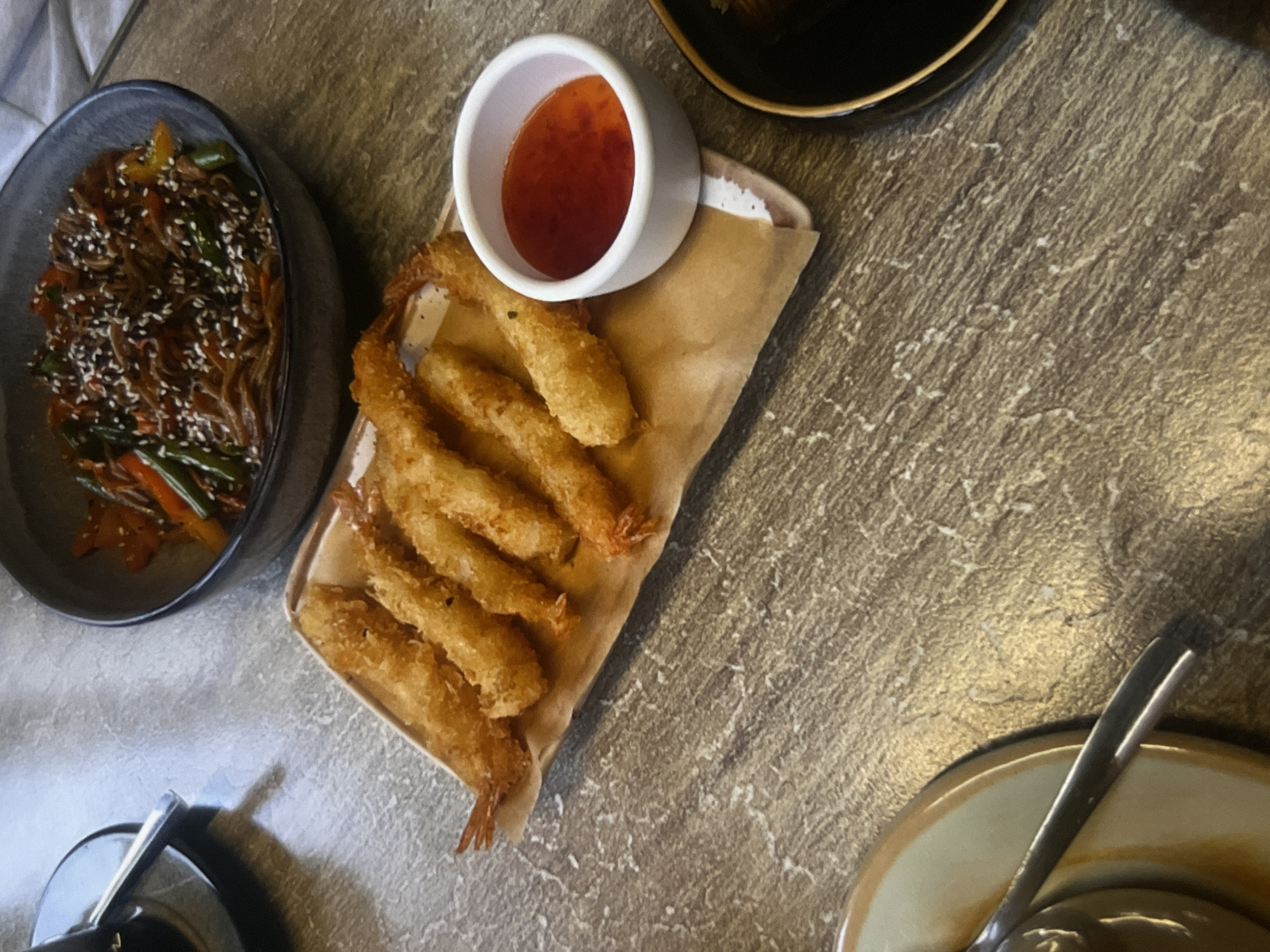 A plate of fried shrimp with a small bowl of dipping sauce sits on a table next to a bowl of noodles. The shrimp are golden brown and arranged in a line on a piece of brown paper. The dipping sauce is red and appears to be sweet chili sauce. The noodles are dark brown and topped with sesame seeds and vegetables. The table is made of wood with a textured surface. A plate with a spoon is visible in the lower left corner of the image. A dark bowl with noodles is in the upper right corner.