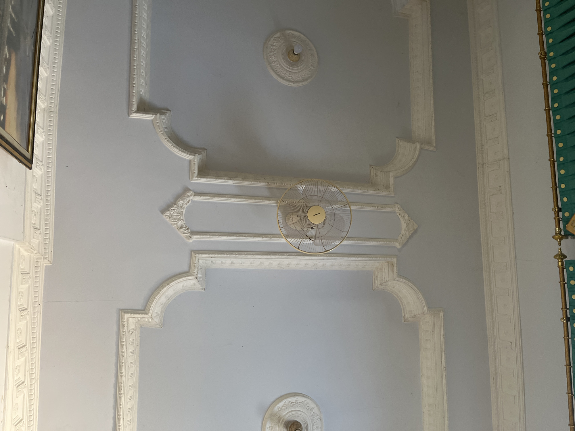 The image shows a close-up, high-angle view of a decorative ceiling in a room. The ceiling is painted a light blue-gray and features elaborate white plasterwork in a neoclassical style. There are recessed panels with intricate molding and a central decorative medallion. A vintage-style oscillating fan, with a gold-colored cage and a beige central plate, is suspended from the ceiling. To the left of the fan, another decorative medallion is visible, and to the right, a similar medallion with a visible light bulb is present. The overall impression is one of ornate interior design. The location in Glagah Kulon, Indonesia, suggests this is likely a domestic or commercial interior space within that region.