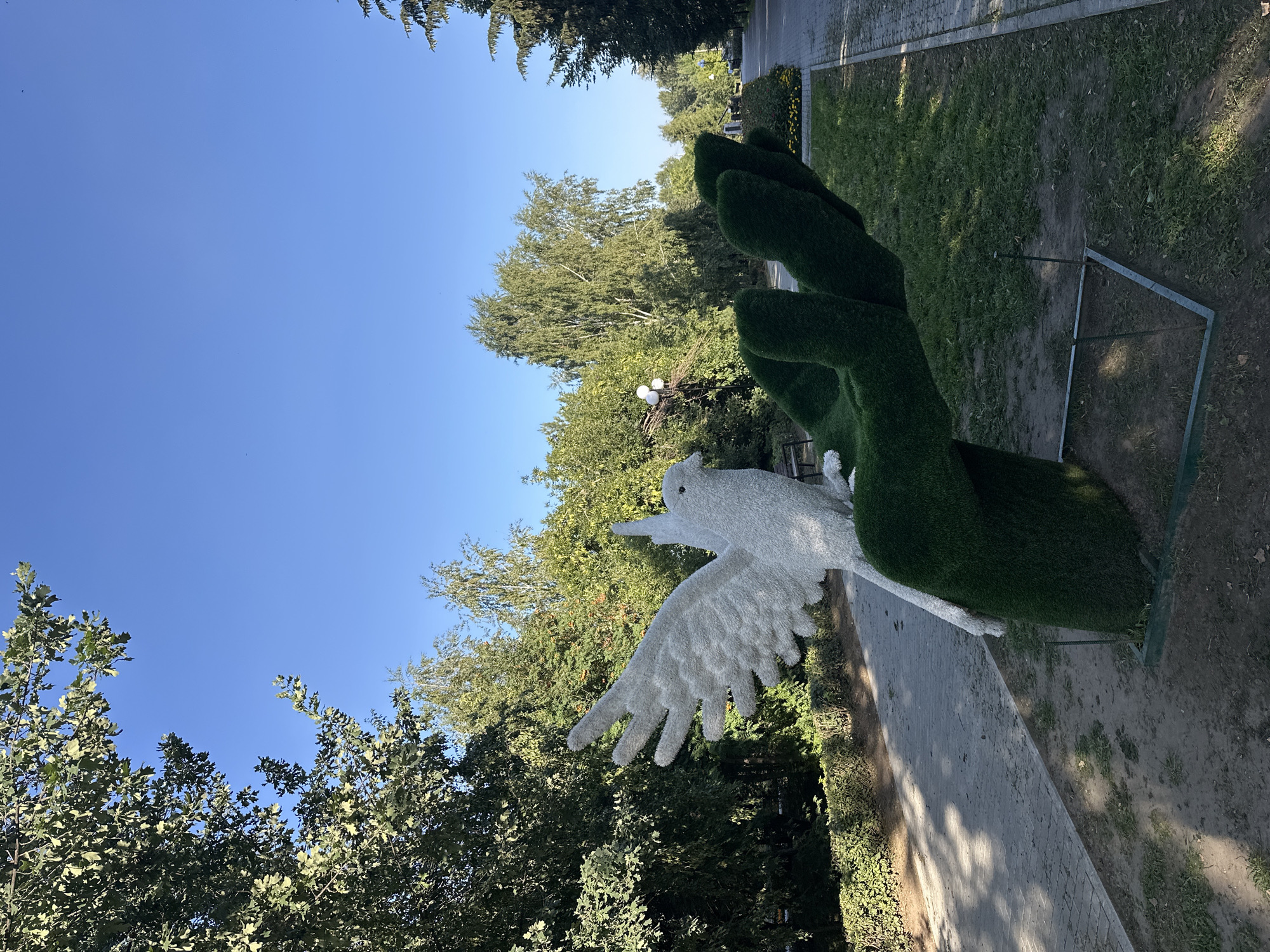 In Nogornyi, Russia, a topiary sculpture of a white dove with outstretched wings rests on a green, hand-shaped base in a park. The sculpture is situated on a dirt and grass area next to a paved walkway. Trees and foliage surround the sculpture, creating a lush, green environment. The sky is a clear, bright blue, indicating a sunny day. The scene is tranquil and appears to be a public space designed for leisure and appreciation of art and nature.