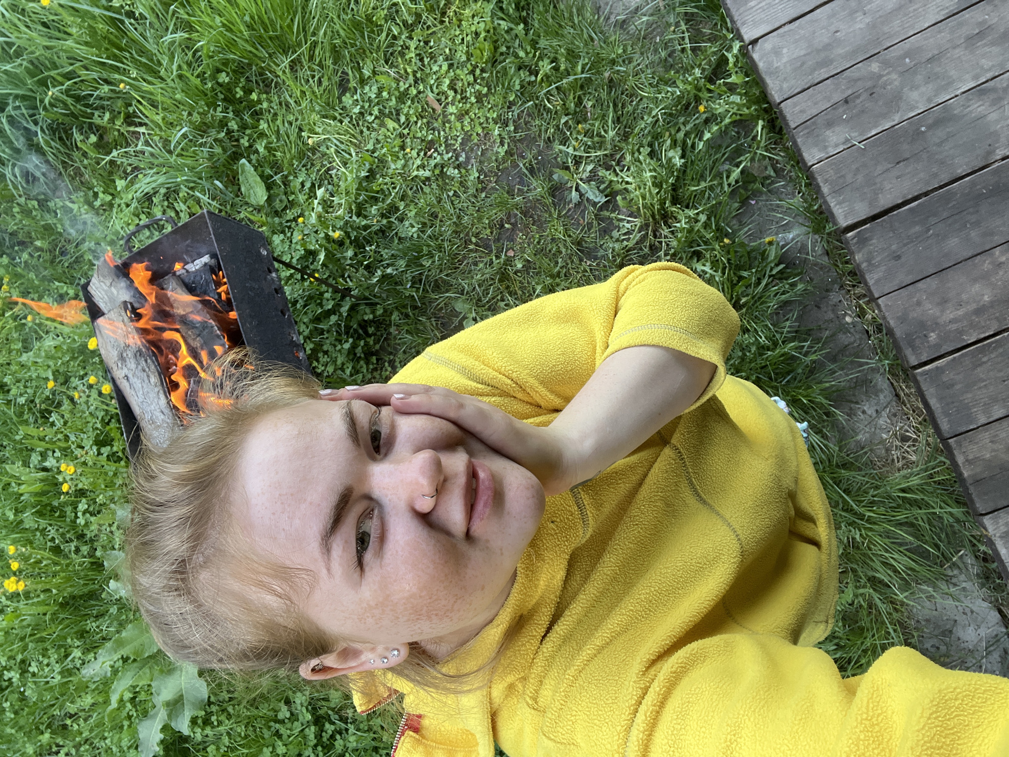 A woman wearing a yellow fleece jacket is positioned in front of a burning grill on a lawn. The grill is filled with wood and has flames emanating from it. To the right of the lawn is a wooden deck. The setting is likely in an outdoor space in or near the city of Chaikovsky, Russia, as per the user's request.