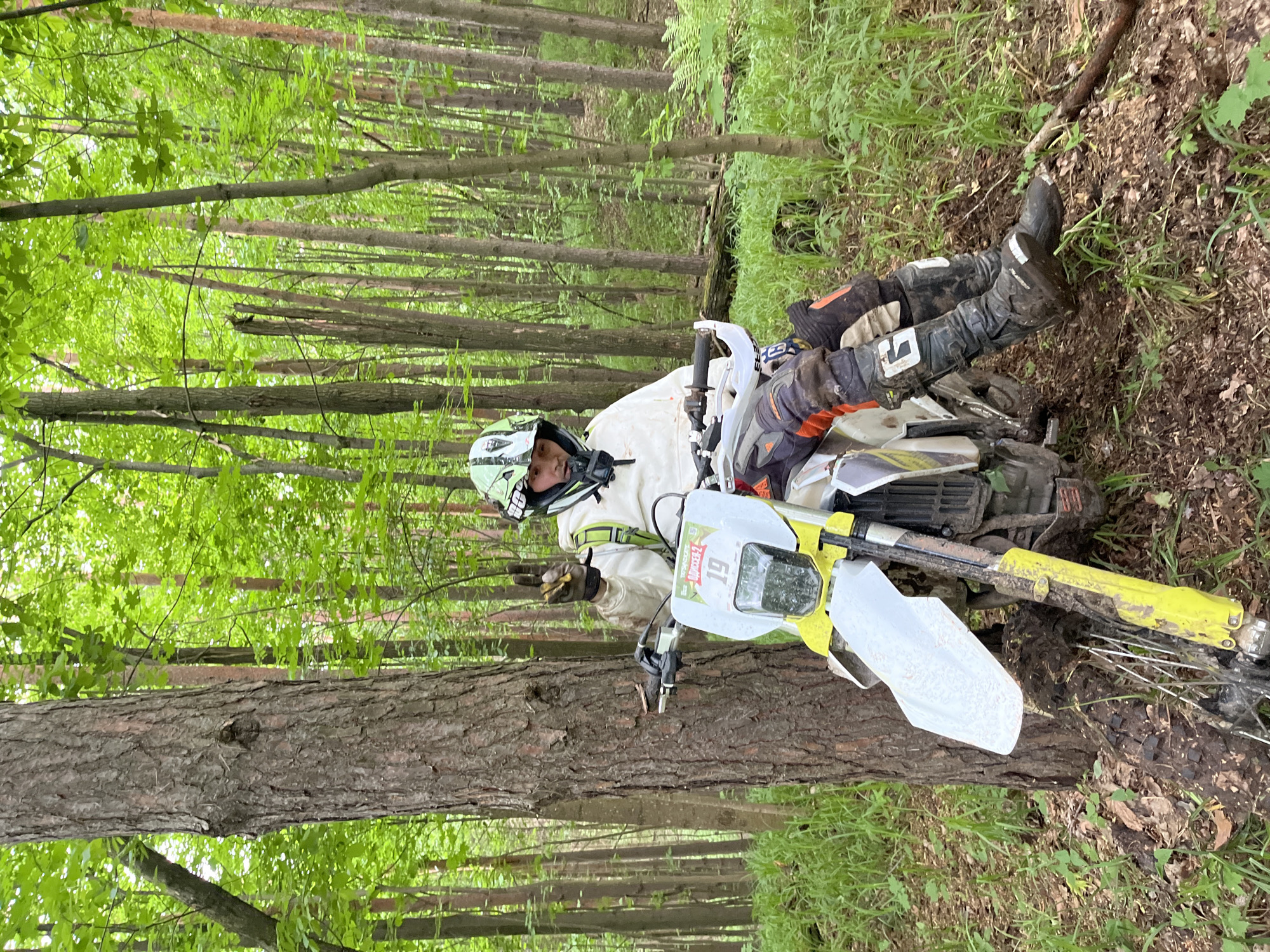 A man wearing a helmet, motocross boots, gloves, and motocross gear is lying on the ground next to a dirt bike. The dirt bike appears to have crashed or fallen over. The scene is a wooded area with trees and greenery. The location is in Gruzinskii, Russia.
