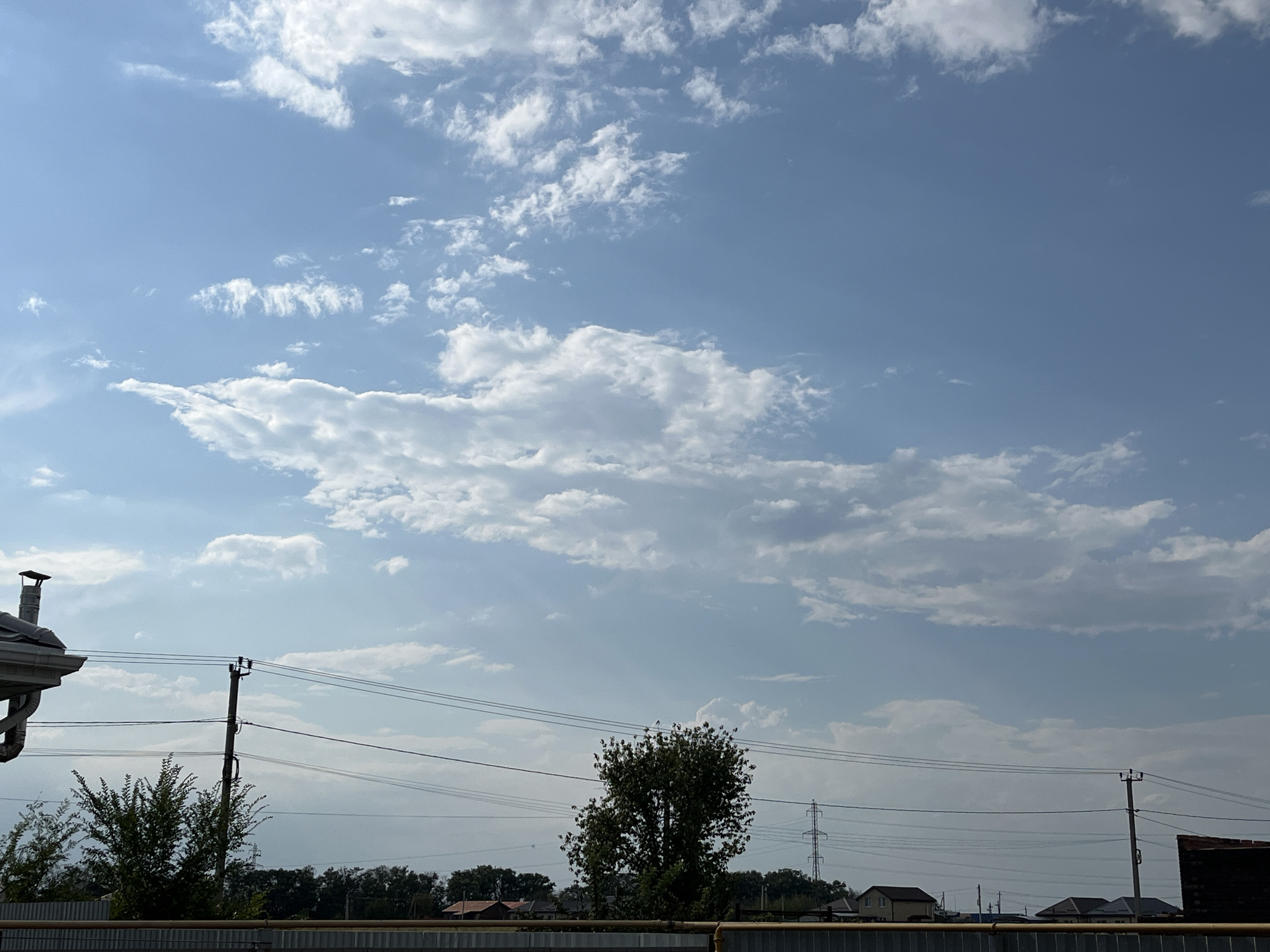The image depicts a sky filled with scattered white cumulus clouds against a clear blue sky. In the foreground, there are power lines strung between utility poles, a residential fence, and the roof of a building. The background shows a line of trees, houses, and a tall industrial tower. The scene appears to be an outdoor, daytime setting in a suburban or rural area. The location is Taganrog City District, Russia.