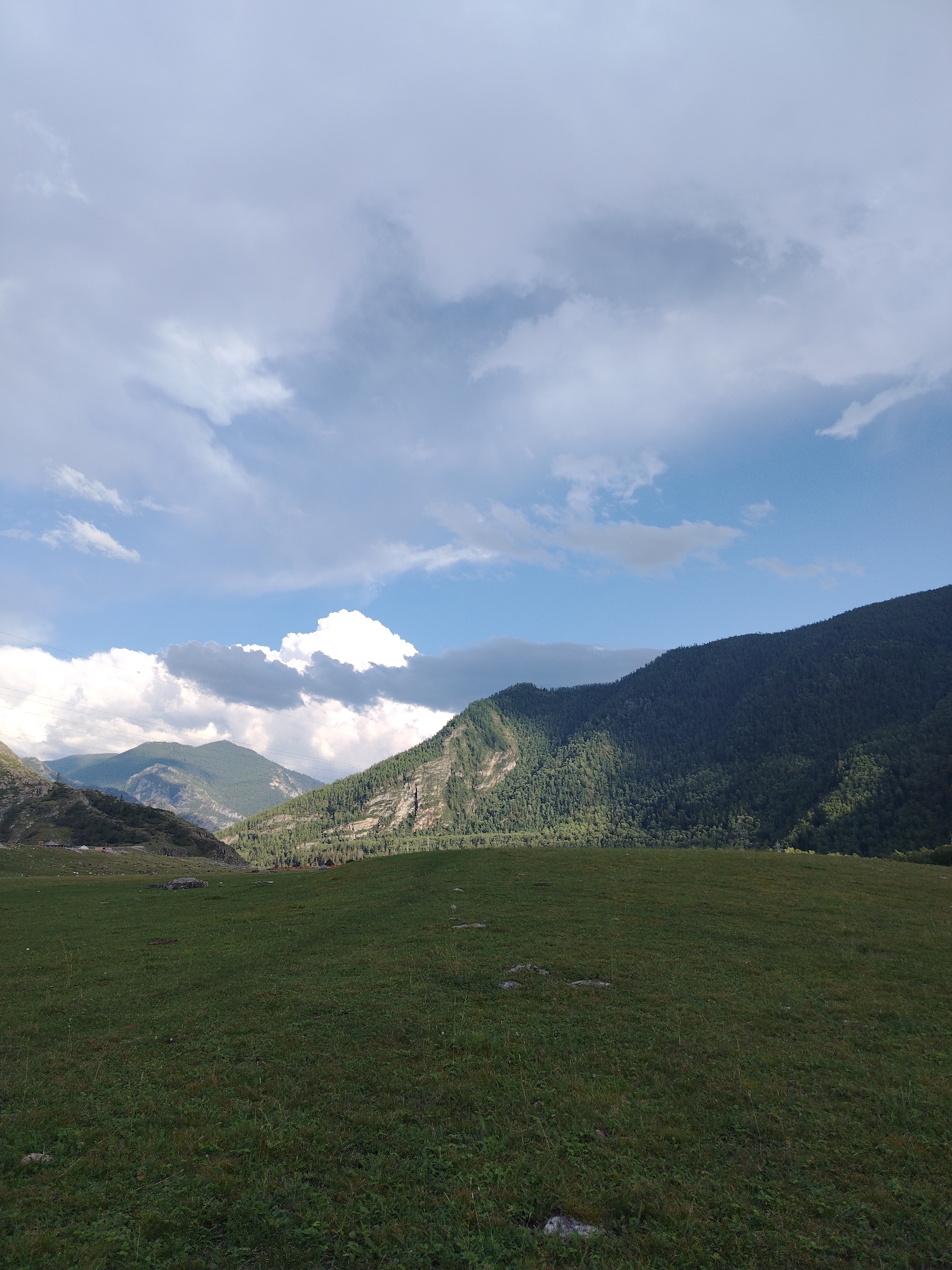 A panoramic view of a verdant valley with steep, tree-covered mountains dominates the scene. The foreground features a gently sloping, grassy field dotted with scattered rocks. In the midground, the valley opens up, revealing a cluster of buildings nestled amongst the trees on the lower slopes of the mountains. In the distance, more mountains rise, their peaks softened by a haze. The sky above is a dramatic mix of bright blue sky and large, white, fluffy clouds, with some darker, more ominous clouds also present, suggesting a dynamic weather system. The lighting indicates it is daytime, with sunlight breaking through the clouds to illuminate parts of the landscape. The overall impression is one of natural beauty and rugged, untamed wilderness in a remote mountainous region. The location is identified as Iodro, Russia.