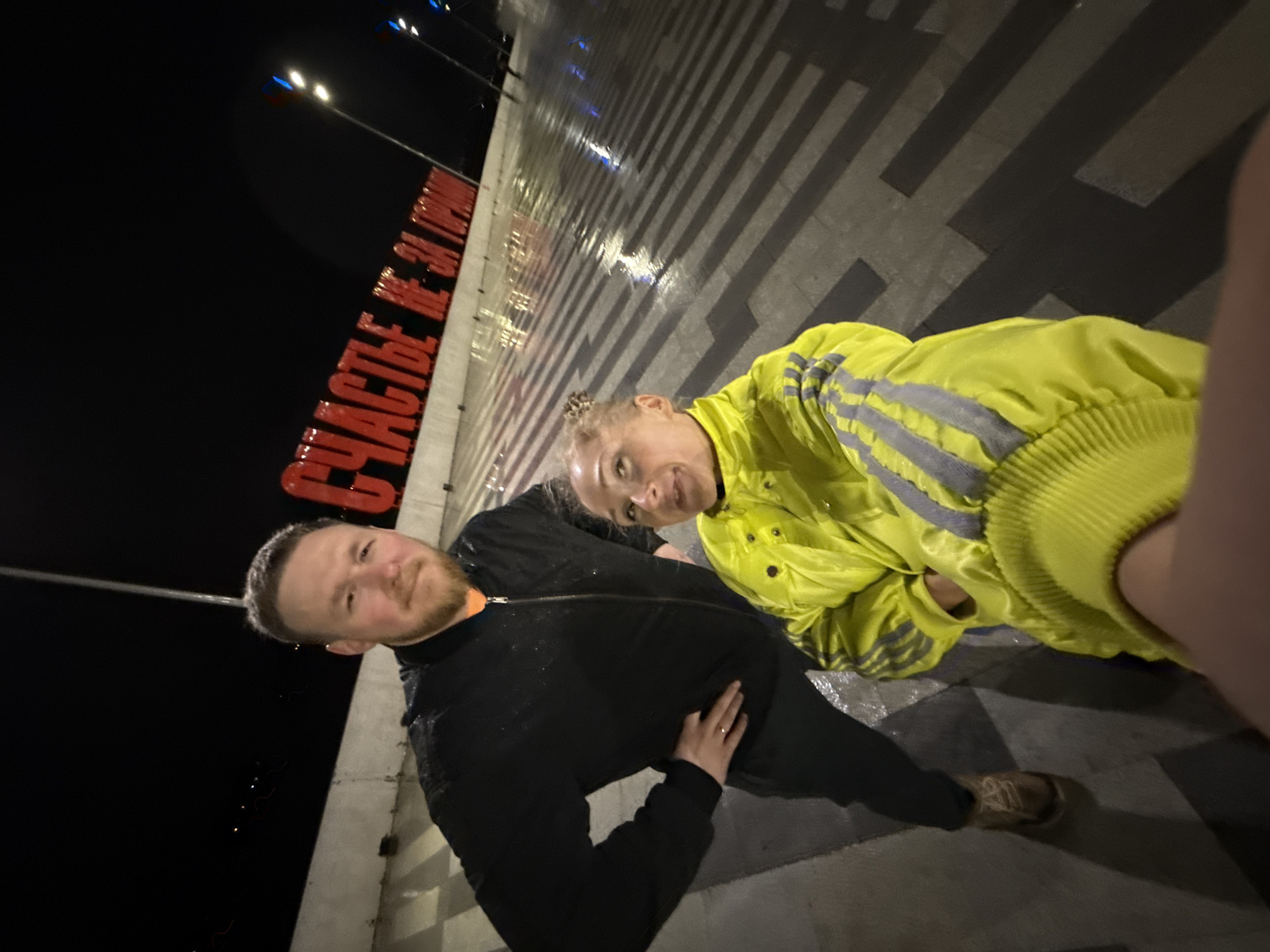 Two individuals pose in the dark, likely at night, in Perm, Russia, near the red illuminated letters spelling out "Счастье не за горами" (Happiness is not far off). A man is dressed in black, and a woman in a bright yellow jacket. The ground is wet and reflective. They are standing on a paved area with contrasting dark and light rectangles. Streetlights illuminate the background.