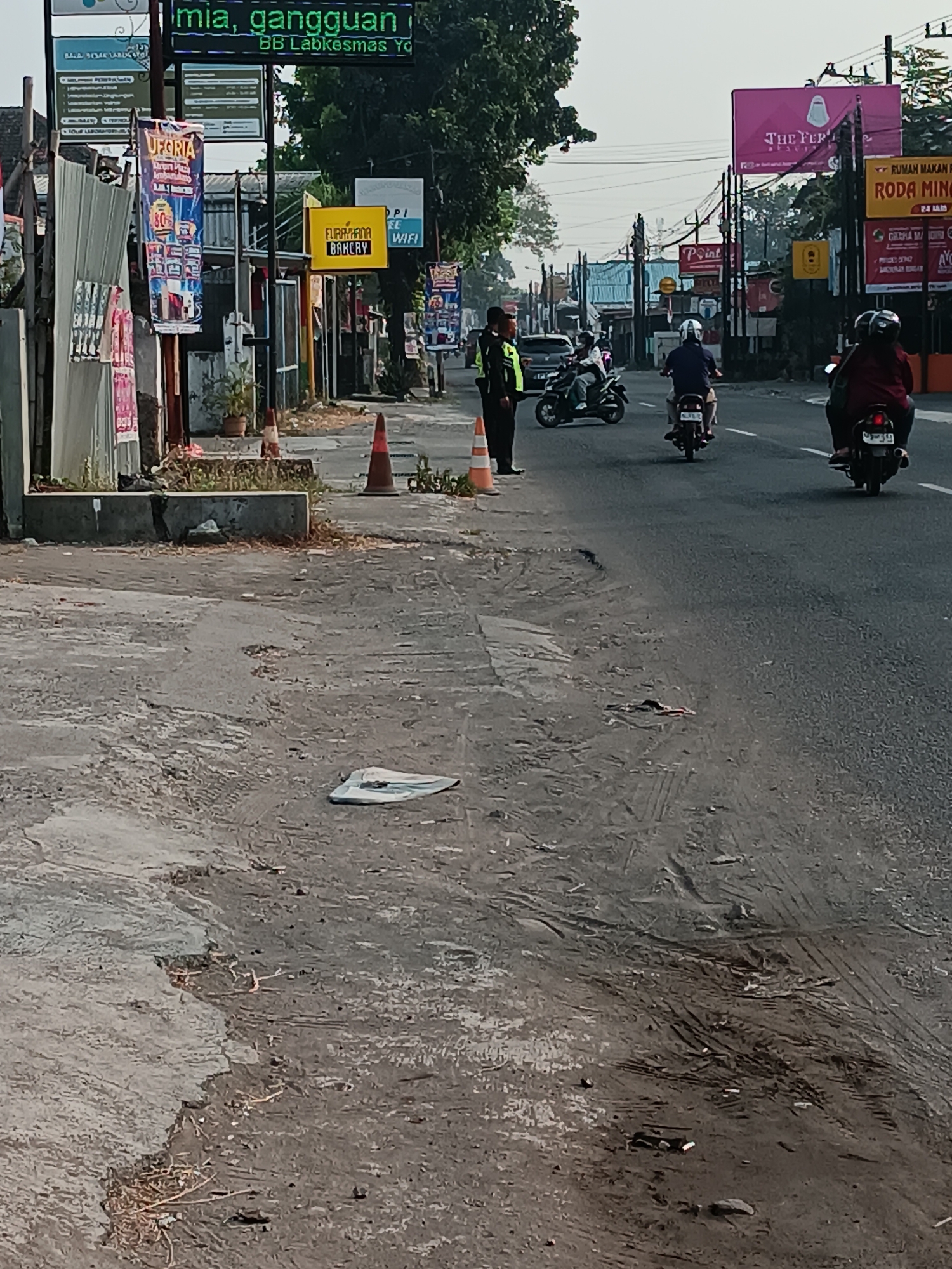 The image captures a street scene in Banguntapan, Indonesia. On the left side of the road, a metallic fence separates the sidewalk from a building. A tall, vertical banner with the word "UFURIA" and "80%" is visible, along with other advertisements. A yellow sign with "FUJIYANA BAKERY" is also present. Two traffic cones are placed on the sidewalk. A person in a yellow and green vest, likely a traffic officer, stands near the cones.
The road itself is paved and appears to be a two-lane street with traffic moving in the same direction. Several motorcycles are on the road. One motorcycle is ridden by a person wearing a dark shirt and helmet. Another motorcycle carries two people, one in a maroon shirt and helmet, and the other in a dark shirt. In the distance, more motorcycles and a car can be seen.
Overhead, utility poles with numerous wires are visible. In the background, buildings and signs, including a large pink billboard with "THE PER...", suggest a commercial area. The sky is overcast. The ground on the side of the road is rough and appears to be a mix of dirt, gravel, and some debris, including a white plastic object resembling a mask or bag. Tire tracks are imprinted on the dirt surface.