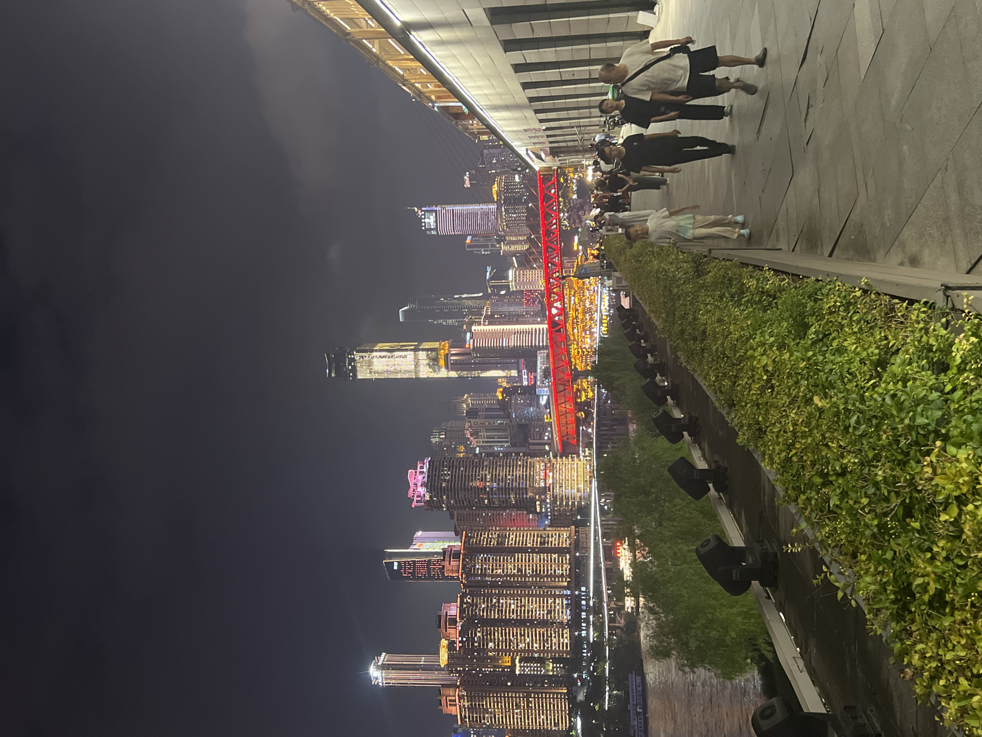 The image depicts a nighttime scene in Jiangbei Cheng Street, China. A wide, paved walkway runs along the right side of the frame, bordered by a low, well-maintained hedge. Several people are walking along the walkway. In the foreground, a young boy in a white t-shirt and light-colored pants walks towards the left. To his right, a man in a black t-shirt and dark pants walks in the same direction. Behind him, another man in a black t-shirt and dark pants walks, followed by a man in a white t-shirt and dark shorts. Further down the walkway, more individuals are visible.
To the left of the walkway, the ground slopes down towards a body of water, likely a river, with a boat illuminated by lights visible on the water. Along the embankment between the walkway and the water, a line of dark spotlights is mounted. Green trees and other vegetation are present along the bank.
In the background, a cityscape is illuminated by a multitude of lights. Numerous high-rise buildings, some with intricate lighting designs, dominate the skyline. A striking red bridge with illuminated arches spans across the water, connecting parts of the city. The overall scene suggests a vibrant urban environment at night, with people enjoying a promenade along the waterfront.