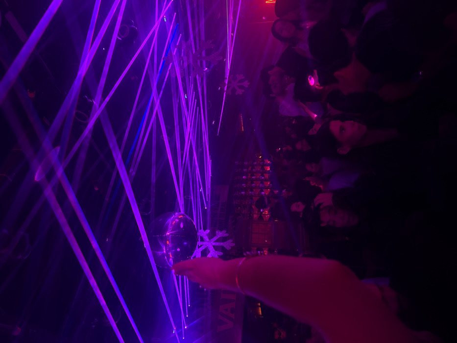 An interior view depicts a crowded nightlife venue in San Bernardino Tlaxcalancingo, Mexico, illuminated predominantly by purple laser beams and dim ambient lighting. The ceiling is crisscrossed with numerous bright purple laser lines, interspersed with some blue light. A reflective disco ball hangs in the upper-left quadrant, with white snowflake-shaped decorations suspended nearby and further into the venue.
In the immediate foreground, an individual's right arm and hand are raised into the upper-left, with the palm facing forward. A thin, reflective bracelet is visible on the wrist. Below and beyond this arm, the venue is densely populated with numerous individuals.
To the left, a bar area is visible in the mid-ground, featuring shelves stacked with illuminated bottles. Above the bar, a partial sign displaying the letters "VAI" in white illumination is discernible. Further right, an indistinct red or orange neon sign is present.
Among the visible crowd, individuals are in various postures. One person with dark hair in the mid-ground-left appears to be touching their head. To their right, another individual with dark hair is observing towards the right. Two individuals further right are positioned closely, appearing to be engaged in conversation. The faces and attire of most individuals are indistinct due to the dim, colored lighting. The scene conveys a busy, social gathering.