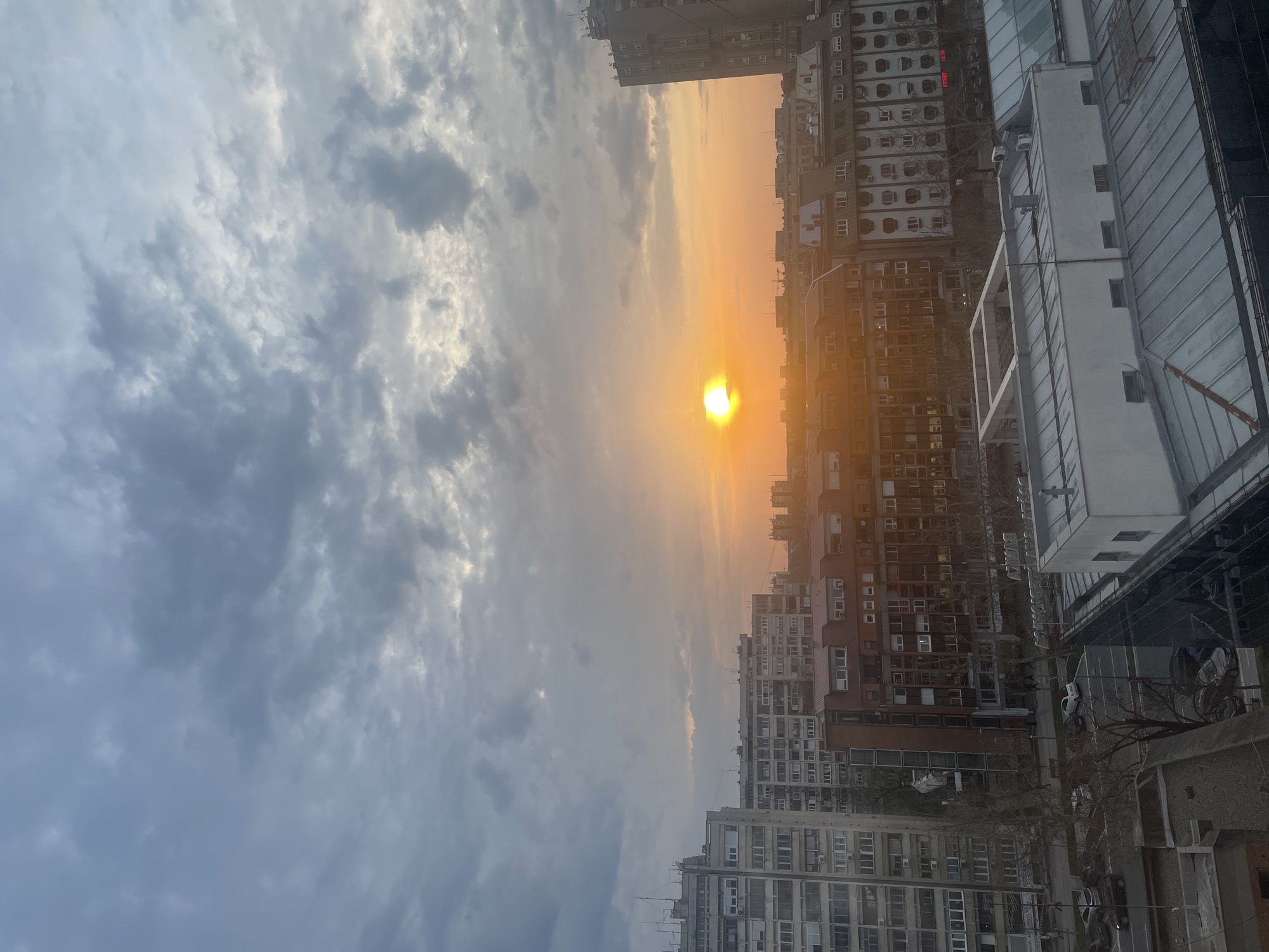 The image shows a cityscape at sunset in Novi Sad, Serbia. The sun is visible, casting an orange glow over the buildings. The sky is cloudy with gray and white formations. Buildings of varying heights and architectural styles dominate the foreground. There are cars parked on the street below. The buildings appear to be residential and commercial structures.