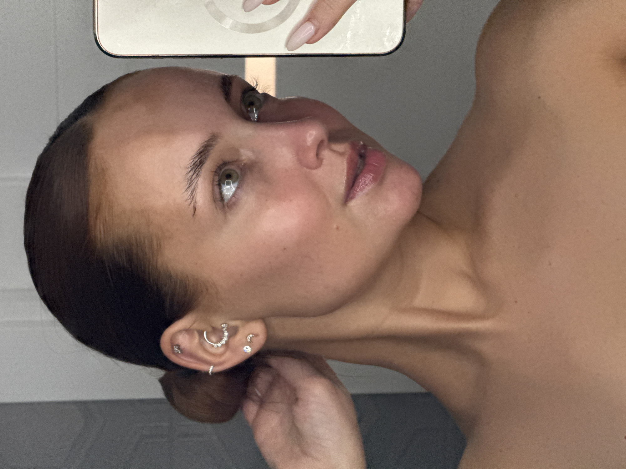 A young woman is taking a selfie indoors. She has light skin, brown hair pulled back, and green eyes. She's wearing minimal makeup. Her left ear is visible with multiple piercings. She's holding a phone in her right hand, partially visible in the top of the frame. The background is indistinct, suggesting an indoor space. The location and city, Chaikovsky, Russia, are not discernible from the image.