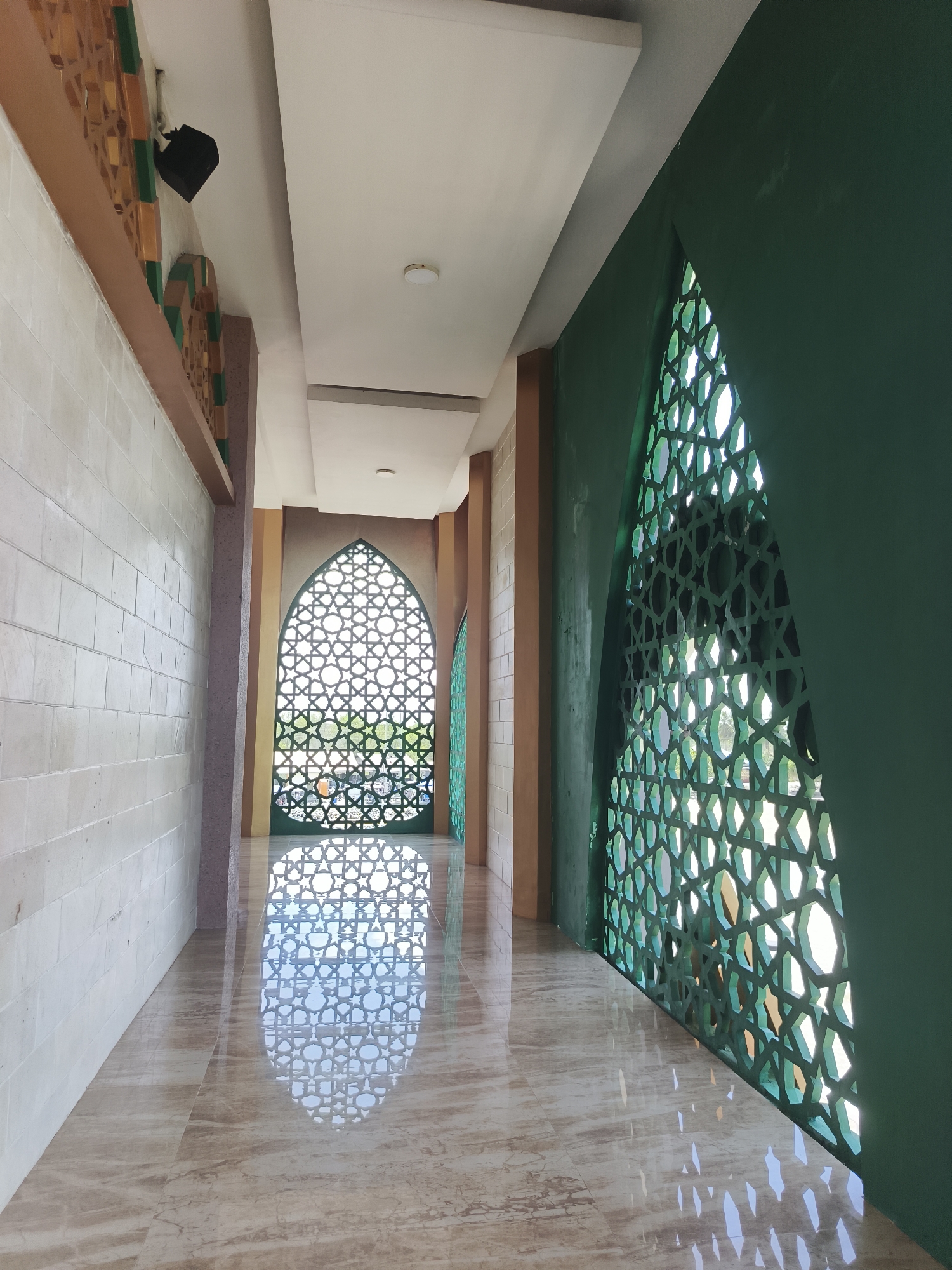 The image captures an interior view of a hallway, likely within a public building, possibly a mosque or cultural center, in Beringin, Indonesia. The hallway features a tiled floor with a polished, reflective surface that mirrors the architectural elements.
On the left, a wall is clad in light-colored rectangular tiles, creating a textured appearance. Above this wall, a decorative frieze in shades of brown and green runs along the top. A black speaker is mounted on the ceiling near this frieze.
The right side of the hallway is dominated by a large, intricate green lattice screen with a geometric Islamic star pattern. Light filters through this screen, casting patterned shadows on the floor.
In the distance, another similar green lattice screen is visible, framing an arched opening that looks out onto a dimly lit outdoor area, possibly a garden or courtyard. The hallway is illuminated by overhead lights, and the overall impression is one of architectural beauty and cultural significance. The scene appears quiet and unoccupied, with no people visible.