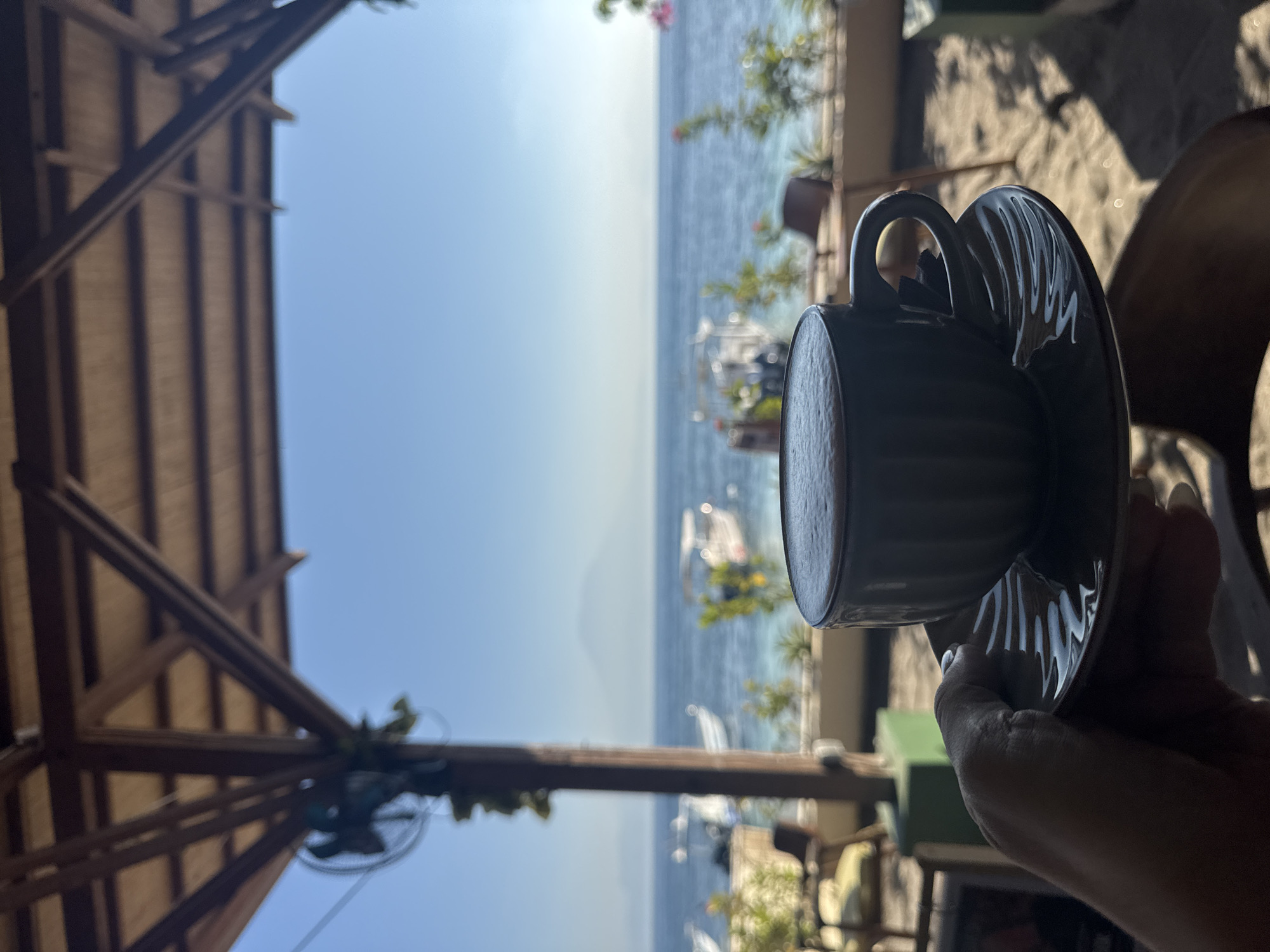 The image captures a first-person perspective from beneath a wooden-beamed and slated canopy structure, looking out over a bright, clear ocean scene. In the lower-center, a hand with light-toned skin and white-painted fingernails holds a dark, ribbed ceramic coffee cup with a matching saucer. The cup contains a beverage topped with a layer of light foam.
The immediate surroundings beyond the hand and cup are partially shadowed but reveal a sandy ground surface with illuminated patches. To the left, a blurred green rectangular base and indistinct furniture are present. To the right, a dark, blurred chair is visible.
The midground features a calm, blue ocean, with areas of lighter turquoise water near the shore. Multiple white and light-colored boats are floating on the water. A line of blurred green foliage and some small pink flowers runs along the coastline, separating the viewing area from the sea.
In the distant background, under a clear, bright blue sky, a large, hazy mountain, characteristic of Mount Agung, is visible on the horizon. This specific geographical feature places the scene in Nusa Penida, Indonesia, looking northwest towards the island of Bali.
The overhead structure consists of dark wooden beams forming a framework, supporting a lighter wooden slatted ceiling. A blurred black fan is attached to a vertical wooden post on the upper left side of the frame. The lighting indicates bright outdoor conditions, contrasting with the shaded interior area where the subject is situated.