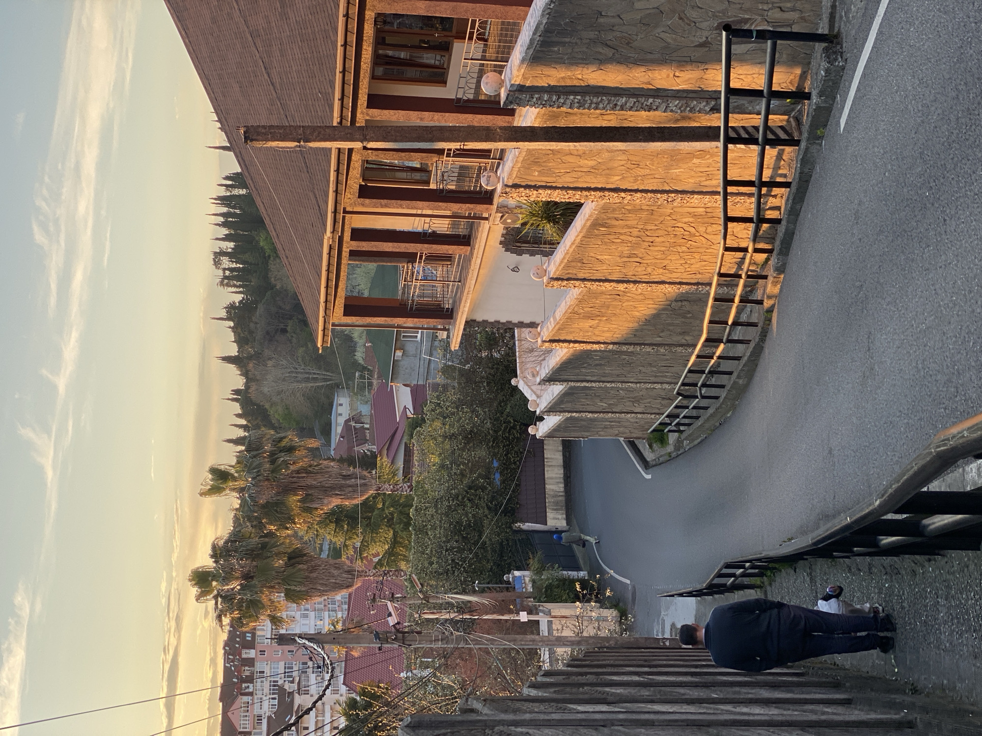 A street scene, likely Sochi, Russia, displays a multi-story building with a brown shingled roof and beige exterior stone facade along a sloping paved road. The building features balconies with dark wood-toned railings. A black metal guardrail runs along the roadside. In the foreground, a man in a dark suit stands on stone steps. In the distance, residential buildings with red roofs are visible amidst green palm trees and other vegetation. The sky is pale with light cloud cover. A person is visible walking along the street in the middle ground. Electrical wires are visible overhead.