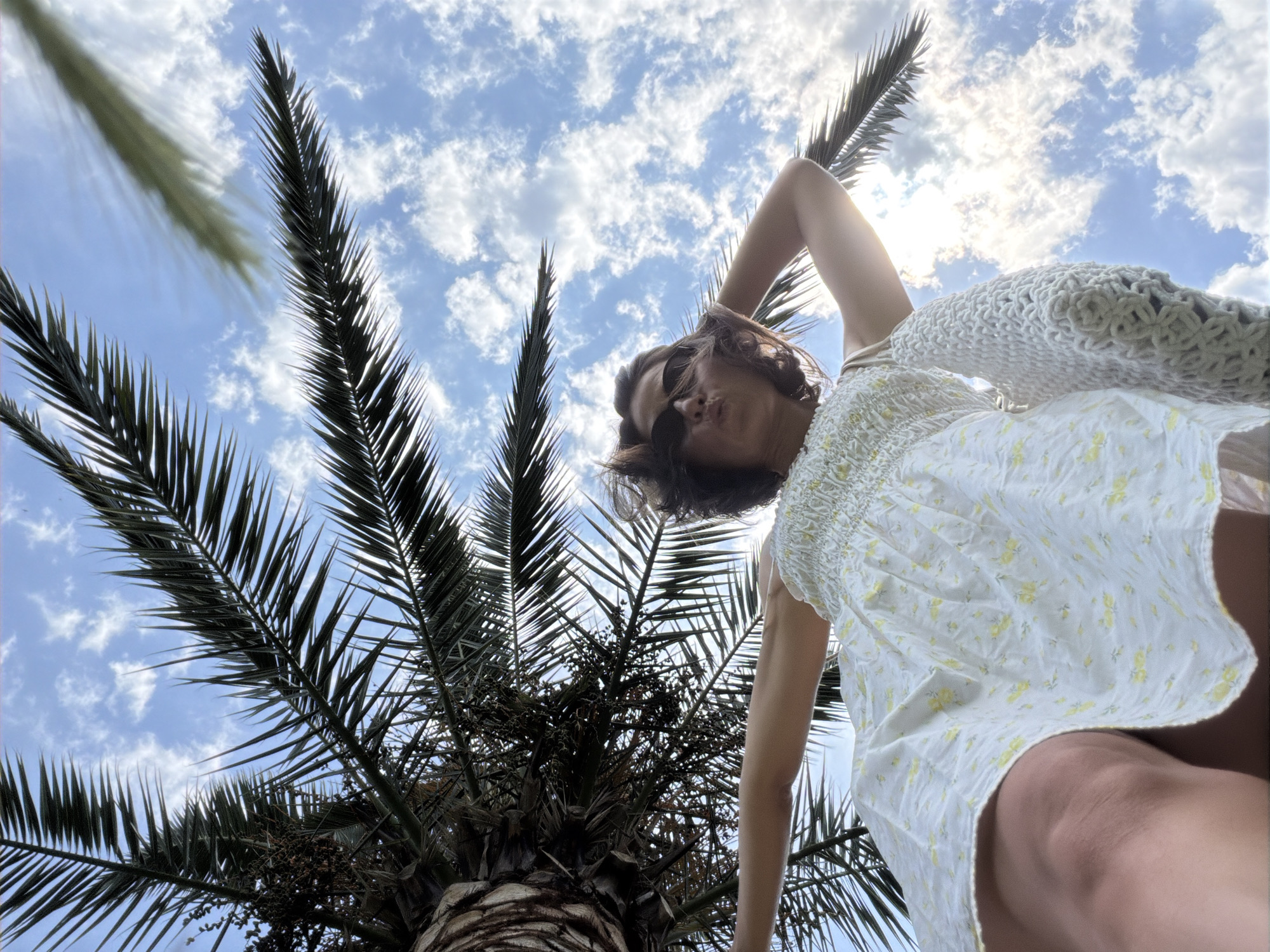 A low-angle, wide shot captures a woman standing outdoors beneath the fronds of a palm tree, with a bright blue sky dotted with white clouds in the background. The woman, with dark hair and sunglasses, wears a white dress with a floral pattern and holds a cream-colored woven bag over her shoulder. She is captured from the chest down, with her right arm raised and her left arm extended outward. The perspective suggests the photograph was taken from a low vantage point, possibly looking up from the ground. The scene is consistent with a warm, sunny day, likely in a tropical or Mediterranean climate, such as the city of Budva, Montenegro, known for its coastal setting and palm trees.