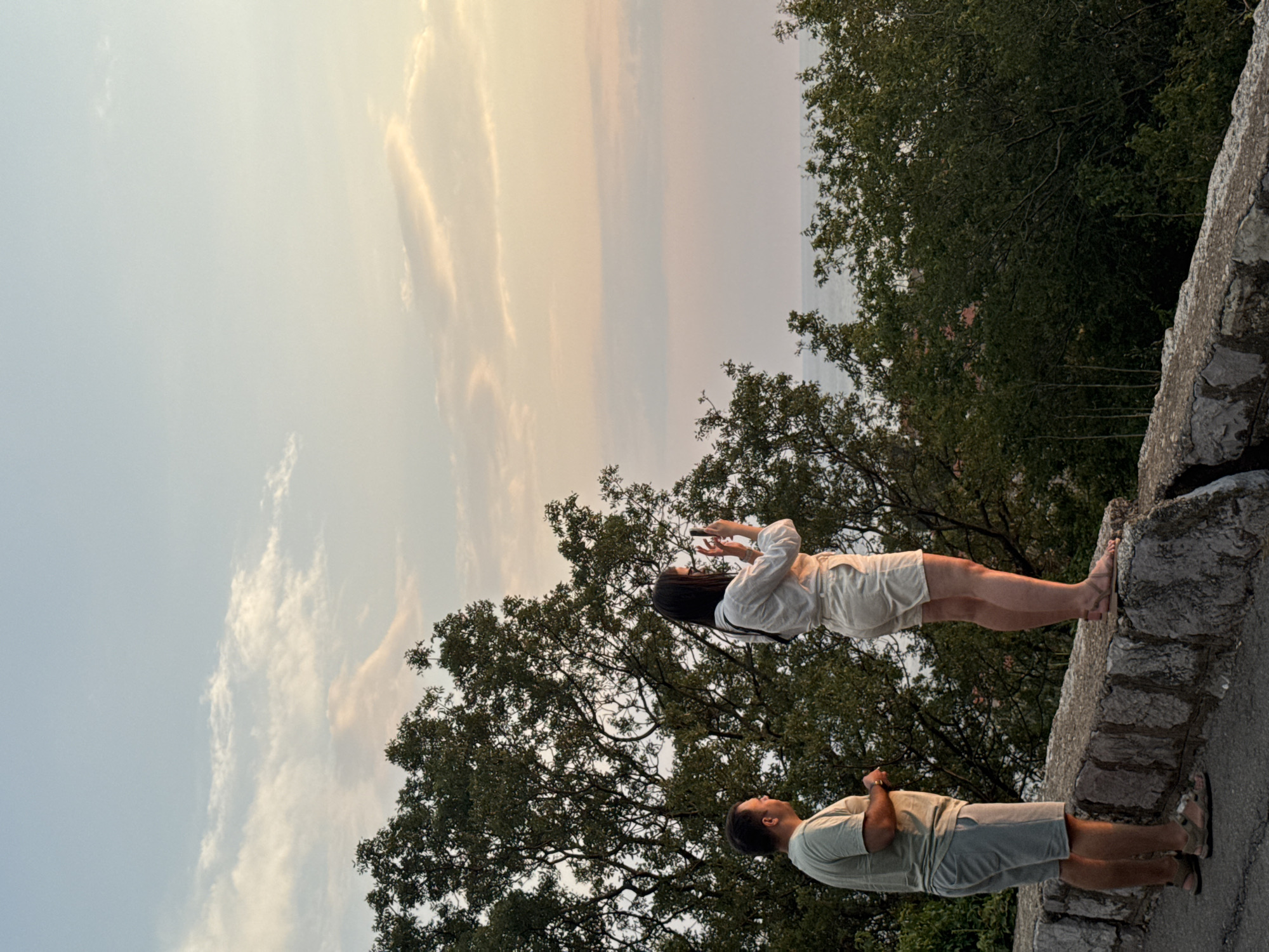 The image depicts a couple standing on a stone wall, possibly overlooking a scenic view. The woman, wearing a white top and light-colored shorts, is holding a smartphone and appears to be taking a picture or recording a video. Her hair is long and dark. The man, in a light olive-colored t-shirt and grey shorts, stands facing away from the camera, looking towards the woman. They are both barefoot, wearing sandals.
The background shows lush green trees and a hazy sky with streaks of orange and yellow, suggesting it is either sunrise or sunset. The sky is partially obscured by clouds. The stone wall they are standing on is rough and uneven, characteristic of an outdoor, possibly historical, location. The overall mood seems peaceful and relaxed, capturing a moment of shared experience or personal reflection.
The image is oriented vertically, and the subjects are positioned on the right side of the frame, with the sky and trees occupying the majority of the left. There is no visible text in the image. The rocky texture of the wall and the dense foliage of the trees provide a naturalistic setting. The faint hints of buildings in the distance, visible through the trees, and the implied coastal landscape suggest a location like Sveti Stefan, Montenegro, known for its picturesque scenery and historic architecture.