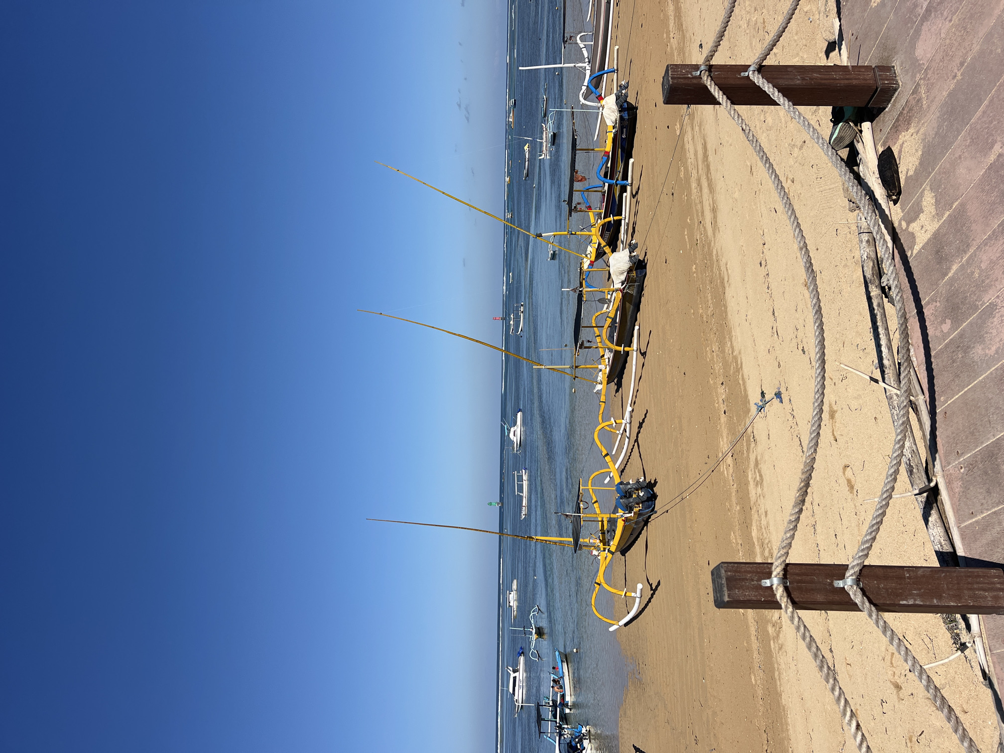 The image depicts a tranquil beach scene in Denpasar, Indonesia, with several traditional outrigger boats lined up on the sand. The boats, primarily yellow and white, are equipped with tall masts and outriggers, characteristic of Indonesian fishing vessels. Some boats have canopies providing shade, and outboard motors are visible on a few.
In the background, the calm ocean stretches out under a clear blue sky, dotted with various other boats, including speedboats and smaller vessels, some anchored and others in motion. A green buoy and a red lighthouse are discernible in the distance, suggesting the presence of navigation aids. The sandy beach slopes gently towards the water, and the foreground is marked by a wooden boardwalk with rope railings, indicating a developed tourist area or recreational spot. The overall impression is one of peaceful coexistence between human activity and the natural beauty of the coastal environment.