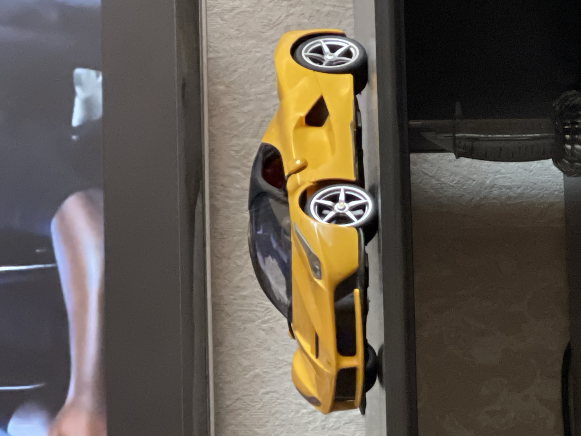 The image captures an indoor scene in Moscow, Russia, primarily featuring a bright yellow model sports car. The car is positioned horizontally on a dark, flat surface, likely a shelf or the top of a piece of furniture, and is oriented with its front facing towards the right of the frame. It displays detailed silver five-spoke wheel rims and a sleek design characteristic of a modern sports car, with a dark tinted windshield and roof section.
To the left of the car and the dark surface, a vertical screen, possibly a television or monitor, is visible. The screen displays content that includes a human leg and a blurry, light-colored, rectangular object against a dark background, suggesting a video or program is being watched.
Further to the right, behind the yellow car and partially obscured by the dark surface, another item is visible. It appears to be a decorative object, possibly a hilt or handle of a sword or a similar ornamental piece, featuring a dark, braided or woven texture. The background wall throughout the image has a light, textured pattern, resembling wallpaper or stucco.
No specific activity is taking place in the immediate physical space beyond the static arrangement of these objects. The time of day, weather, and any emotions are not discernible. There is no visible text in the image.