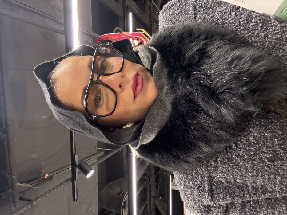 A forensic examination of the image reveals a close-up, chest-up portrait of an adult female, positioned in an indoor commercial establishment within Paris, France. The subject wears a dark grey fabric hood, large tortoiseshell-framed aviator-style glasses with light-tinted lenses, and dark pink lipstick. A grey fabric scarf and a prominent dark grey faux fur collar are draped around her neck. Her outer garment is a textured grey bouclé-knit coat or jacket. A brown patterned strap, possibly from a handbag, is visible on her right side. The woman looks directly forward, seemingly at the camera. The background consists of a dark ceiling with exposed industrial track lighting fixtures and linear LED strips. A partial reflection of an indistinct form is visible in the upper left background. To the far right, a glimpse of a red/pink decorative object or display is present. A white label or document with partial text is visible in the bottom right corner.
