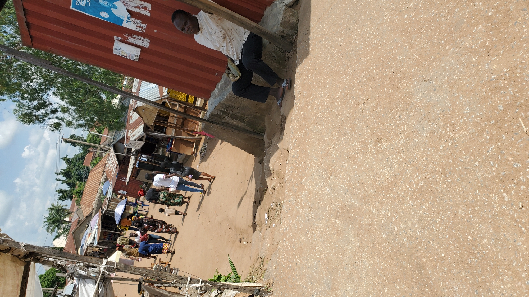In Jalingo, Nigeria, a bustling market scene unfolds on a dusty, uneven dirt road. The foreground is dominated by the textured surface of the road, its tan and gray tones speckled with lighter stones and scattered debris. In the mid-ground, a group of people walk along the road, their figures casting shadows on the ground. A man in a white collared shirt and dark pants sits on a concrete ledge against a large, corrugated red metal structure, leaning against a wooden pole. He holds a light-colored bag on his lap. Behind him, a poster is attached to the red structure. To the left of the road, market stalls with corrugated metal roofs line the street, some with open fronts revealing their wares. The background shows more market structures, trees, and a few residential buildings with similar tin roofs, under a bright, cloudy sky. The overall impression is of a vibrant, open-air marketplace on a sunny day.