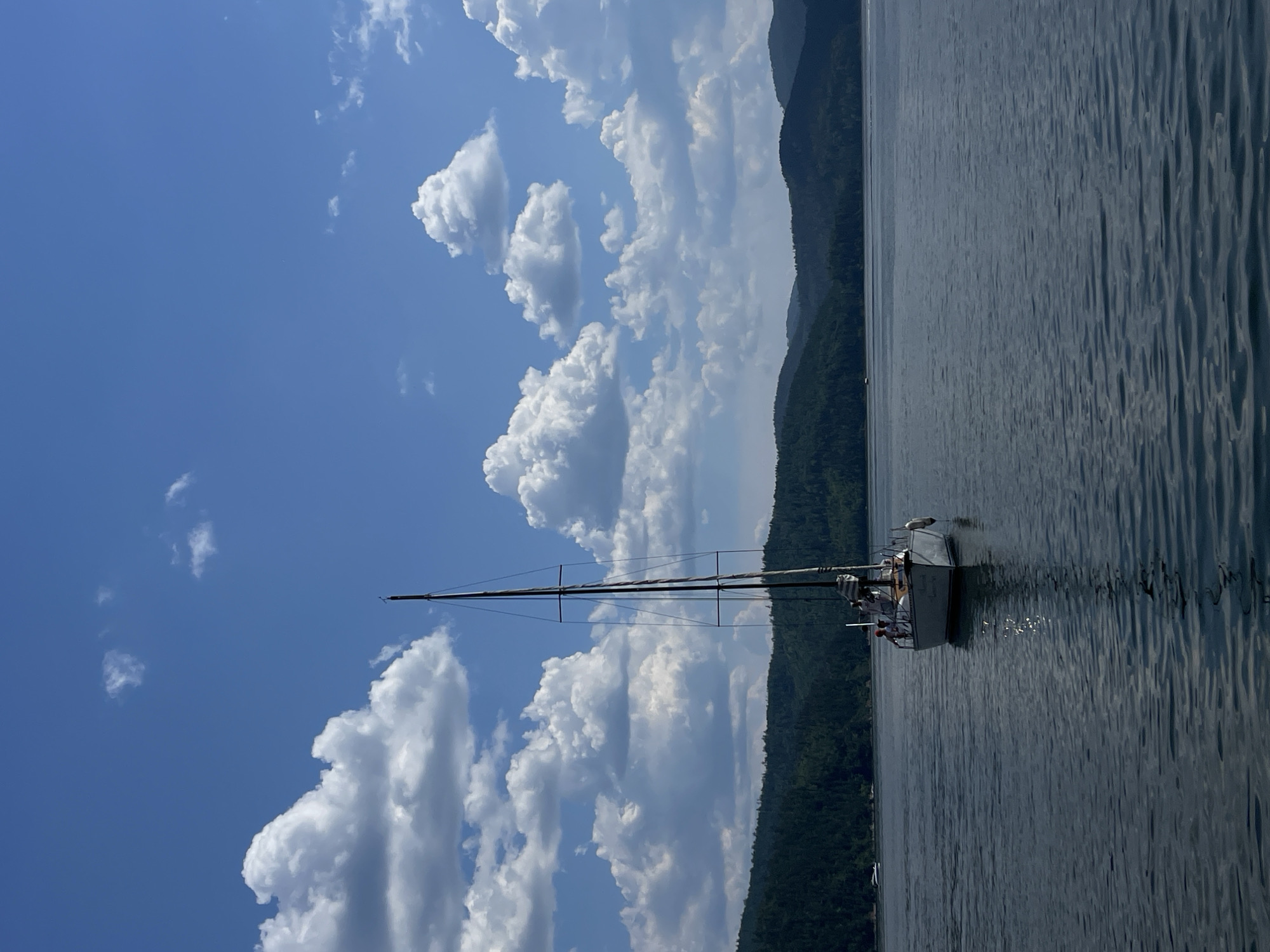 A sailboat with several people on board glides across a calm body of water, with a lush, tree-covered mountainside receding into the background. The sky is a vibrant blue, dotted with fluffy white cumulus clouds, suggesting a pleasant day with fair weather. The sun appears to be high in the sky, casting shimmering reflections on the water's surface. The scene evokes a sense of tranquility and leisure, as the boat seems to be enjoying a serene journey. The image is oriented vertically, with the water and mountains on the right and the sky and clouds on the left. No visible text is present in the image.
