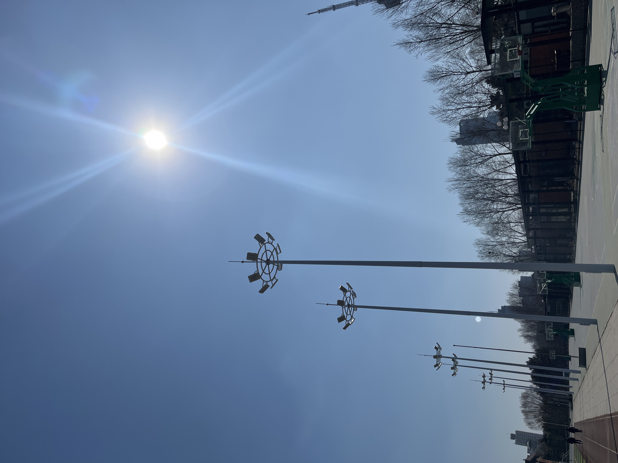 The image depicts an outdoor basketball court on a sunny day in Changchun City, China. The sky is blue with the sun visible in the upper left corner. Several tall light poles line one side of the court. There are basketball hoops, buildings and trees in the background. A communication tower is barely visible.