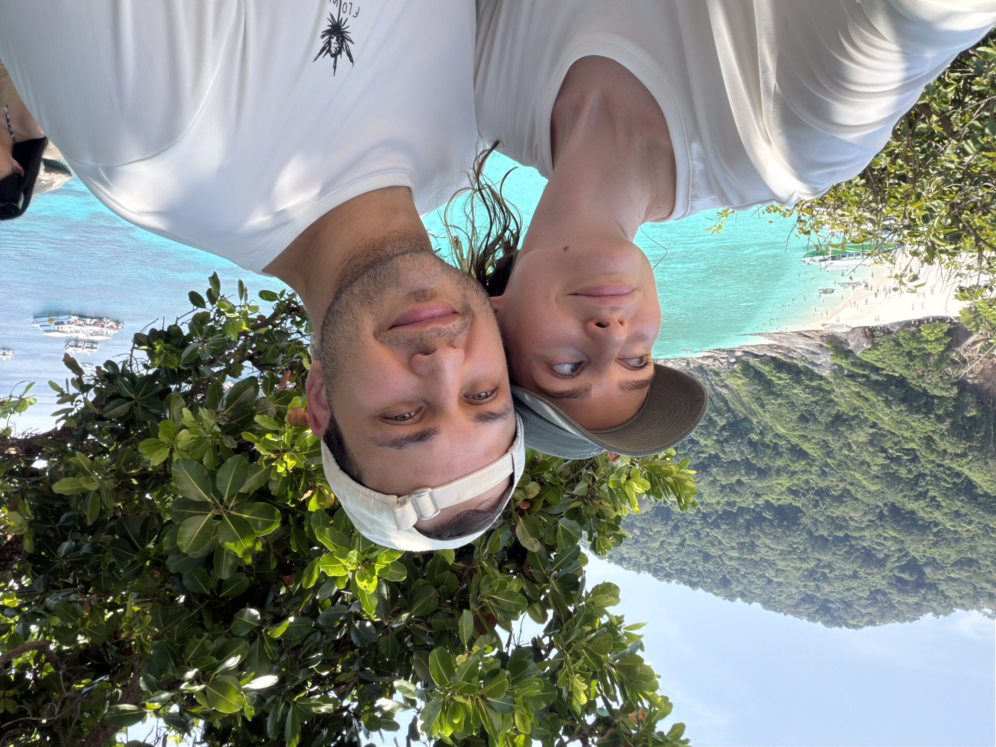 A close-up photograph captures two individuals, one female and one male, positioned in the foreground outdoors. The female, on the left, wears a dark green baseball cap and a light-colored, loose-fitting top, her gaze directed upwards and to the left. The male, on the right, wears a light-colored baseball cap reversed and a white short-sleeved t-shirt with a dark stylized graphic on the left chest, looking directly at the camera. Both individuals have light skin tones; the male exhibits light facial stubble. Partial green foliage from an overhead tree is visible in the upper right. The background features a wide expanse of clear blue sky above a large, densely forested green hill or island. Below the terrain, a white sandy beach is visible, occupied by numerous small human figures. Multiple boats, including smaller vessels near the shore and larger ships further out, are present in the clear turquoise and deeper blue ocean waters. The scene is located in the city of Lam Kaen, Thailand.