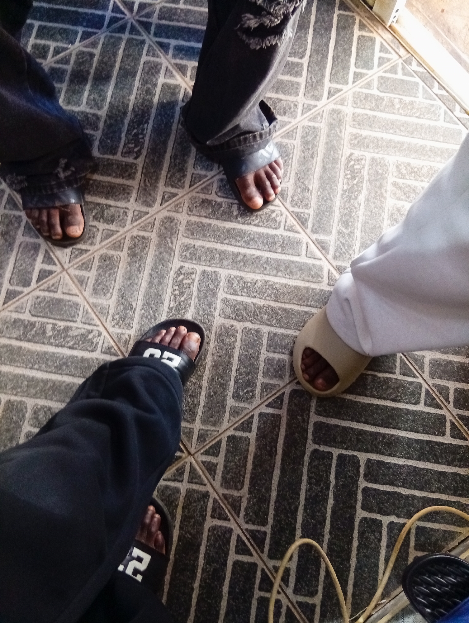 The image shows the lower legs and feet of at least three individuals standing on a tiled floor in Bukuru, Nigeria. One person on the left is wearing dark pants and black slide sandals, with their left foot visible and adorned with a black sandal with white lettering. Another person, partially visible in the upper left, wears dark, distressed jeans and black sandals, with their right foot showing. The third person, on the right, is wearing light-colored pants and tan slide sandals. The tiled floor has a pattern of rectangular grey tiles with darker grout lines. A light-colored object, possibly a wall or door frame, is visible along the top right edge of the image. A yellow cord and a black object are visible in the lower right corner. The scene appears to be an indoor or covered outdoor area.
