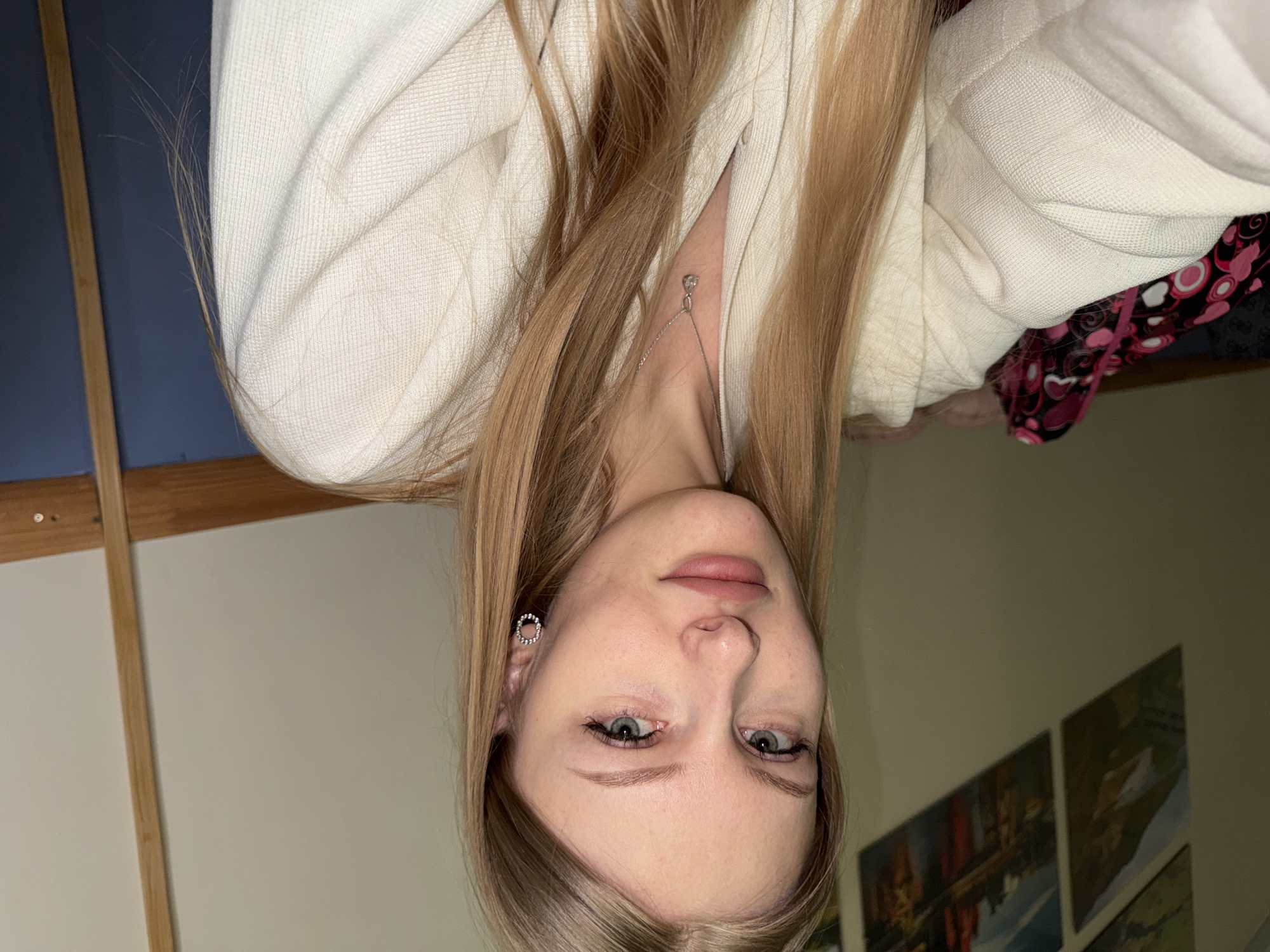 A close-up portrait shows a young woman looking directly at the camera, taken indoors in Perm, Russia. She has fair skin, light-colored eyes, and long, straight, light brown to blonde hair. Her lips have a subtle pink tint. She wears delicate eyeliner and mascara. On her visible left ear, she has a small, circular, silver-toned earring. A thin silver necklace with a tiny, sparkling pendant rests against her chest, partially obscured by her hair and clothing.
She is wearing a light-colored, possibly off-white or cream, textured cardigan or jacket. Peeking out from beneath the cardigan on her right side (viewer's left) is a garment with a black background featuring a pattern of pink and red hearts, circles, and swirls.
The background on the left side of the image (when oriented correctly) features a section of a dark blue wall, separated by a vertical wooden beam from a lighter, possibly beige or off-white wall section. On the right side of the background, several framed pictures or canvases with abstract or landscape art are visible on a light-colored wall. The lighting suggests an indoor setting, likely with artificial light, and the overall mood is calm and neutral. There is no visible text in the image.