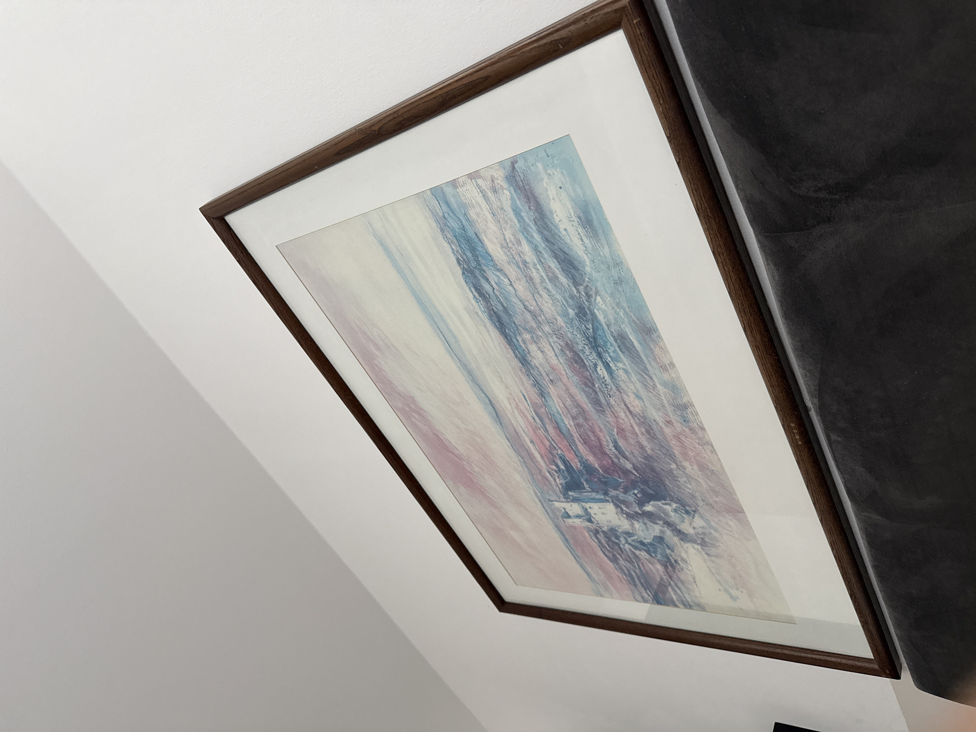 This is a close-up, angled shot of a framed painting hanging on a white wall. The painting depicts a seascape with a lighthouse on a rocky outcrop. The water is rendered in abstract brushstrokes of blue, pink, and purple, suggesting movement and light. The sky is a pale pink and blue. The frame is a dark wood. Below the painting, a dark gray, upholstered surface, likely a piece of furniture, is visible. The overall impression is one of a domestic interior, possibly a living room or hallway. There is no information in the image to indicate its location in Berlin, Germany.