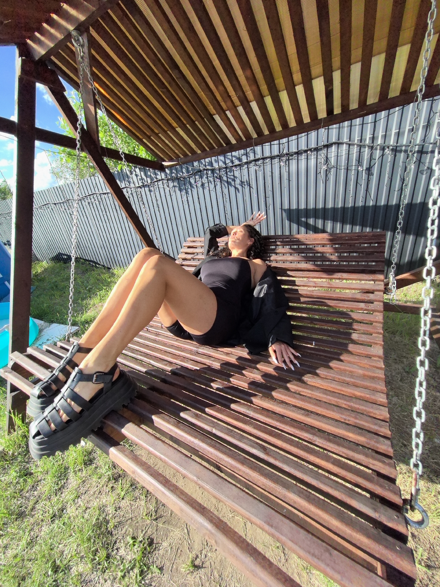 A young woman is lounging on a wooden swing in a backyard setting. She is wearing a black swimsuit, a black jacket draped over her shoulders, and black platform sandals. Her eyes are closed and her head is tilted back, suggesting relaxation or enjoyment of the sunlight. Her legs are extended and her arms are resting on the swing. The swing is suspended by metal chains and has a wooden slatted seat and back. Above the swing, a wooden slatted roof provides shade, casting striped shadows on the seat and the woman.
The scene appears to be outdoors on a sunny day, with a blue sky and green grass visible. A corrugated metal fence runs along the background, with some trees and foliage peeking over it. String lights are attached to the fence. The overall impression is one of a relaxed, warm-weather moment in a residential outdoor space. The time of day is likely afternoon, given the angle of the sunlight and the shadows. There is no visible text in the image. The location context is Chaikovsky, Russia.