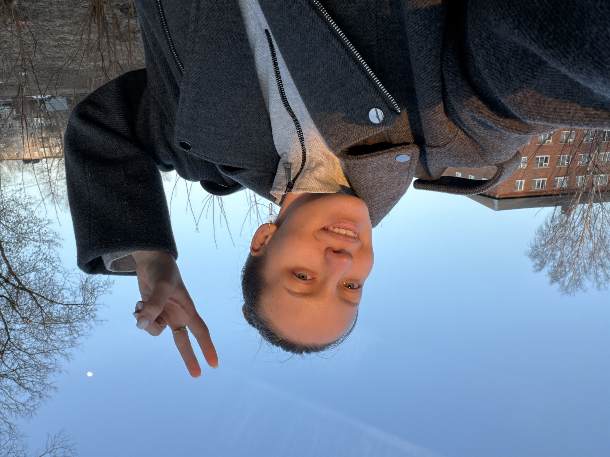 A person, likely female, is seen smiling and holding up a peace sign with her left hand in front of a clear blue sky. She is wearing a grey coat with a visible zipper and a light-colored top underneath. In the background, there are bare trees, a brick building with windows, and what appears to be a residential area. Based on the prompt, the location is Reshetnikovo, Russia.