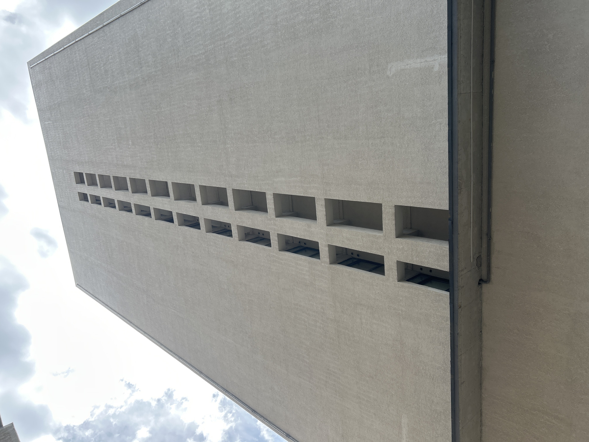 The image shows a tall, gray building with a series of rectangular window openings. The building's exterior appears to be made of concrete. The perspective is from below, looking up at the building. The sky is visible in the background, with scattered clouds. The image is taken in Tbilisi, Georgia.