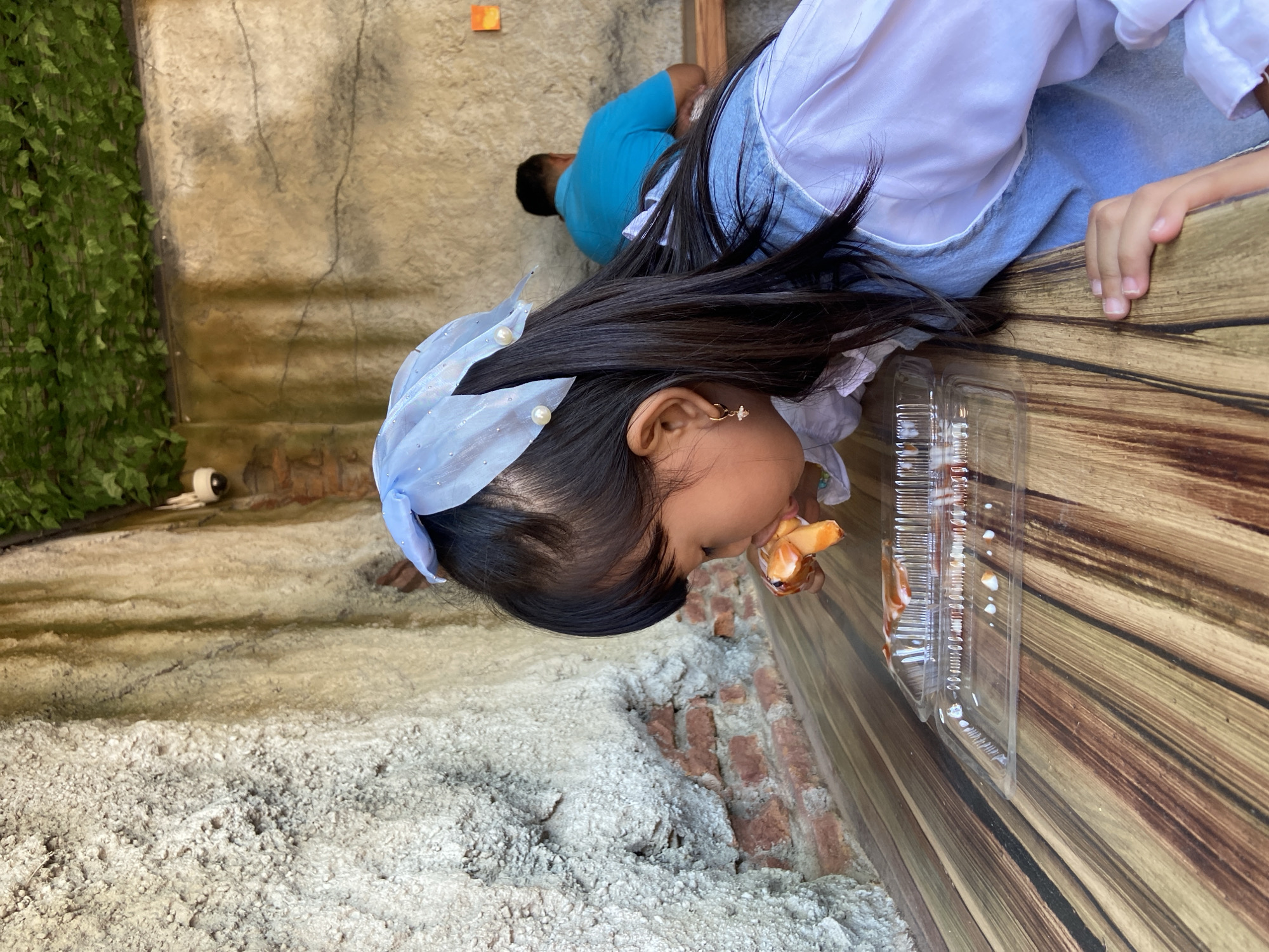 A young girl with long black hair, wearing a blue head covering, a white top, and a denim-colored garment, is eating food from a clear plastic container. She is positioned next to a wooden surface and a textured wall. A small portion of another person wearing a blue shirt can be seen in the background. The background also includes a brick-like wall and greenery. The location is Mangli, Indonesia.