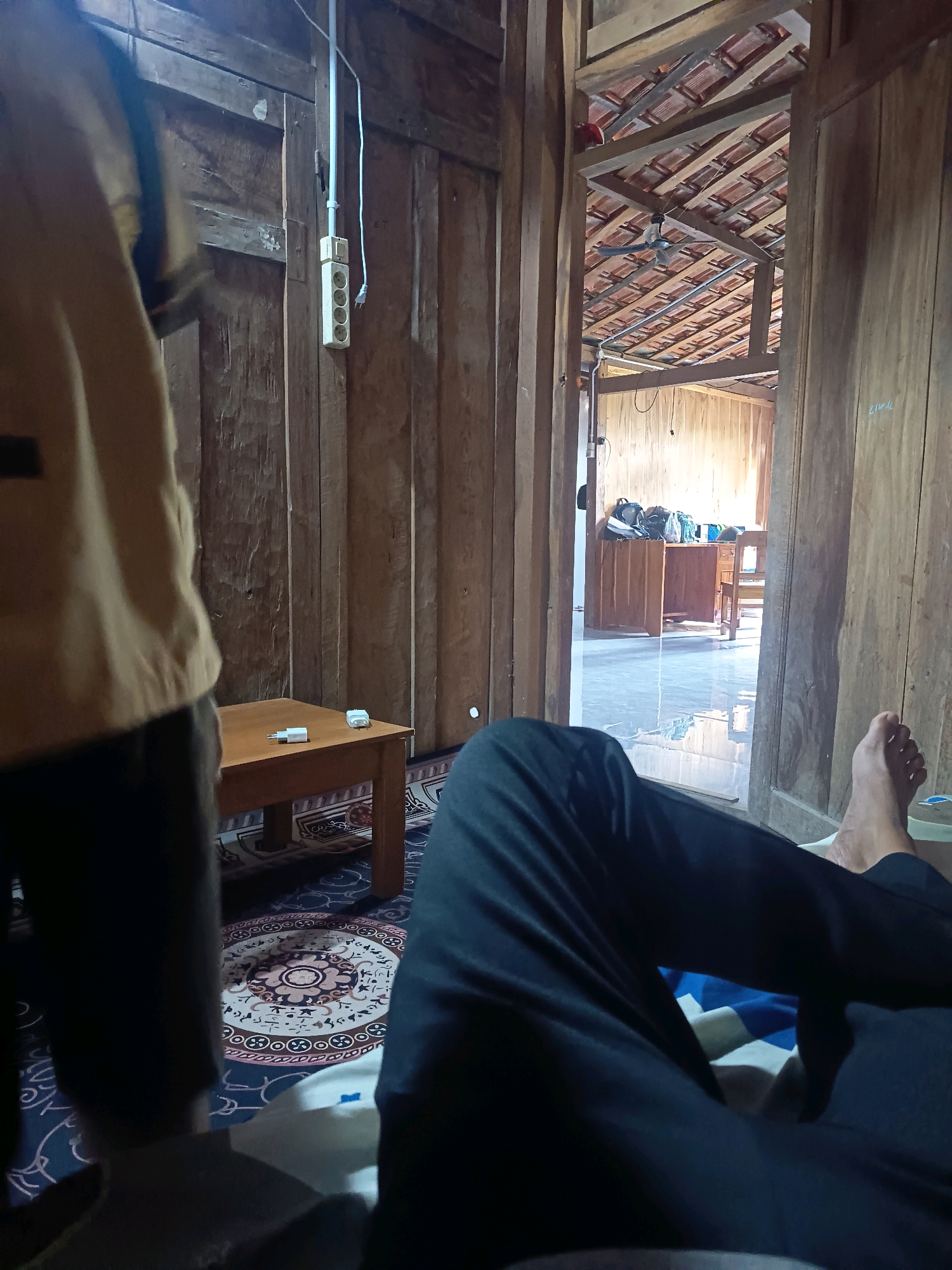 The image depicts an interior scene of a room with wooden walls and a visible ceiling structure of wooden beams and terracotta tiles. In the foreground, a person's legs in dark trousers are visible, resting on what appears to be a bed with blue and white striped bedding. To the left, another person is partially visible from the back, wearing a tan or mustard-colored top and dark trousers. They are standing near a small wooden table with a white electrical adapter on it.
The room has a rustic, somewhat unfinished aesthetic, consistent with construction or renovation. Electrical outlets are visible on the wooden walls, including a multi-outlet strip. In the background, a doorway opens into another part of the house, revealing a desk, chairs, and bags, suggesting it might be a workspace or study area. A ceiling fan is visible in the distant room. The floor appears to be tiled. The lighting suggests it is daytime, with light coming from the open doorway and possibly other sources not fully visible. The overall impression is of a lived-in space, possibly in a rural or semi-rural setting in Sidorejo, Indonesia, given the architectural style and typical materials used in such regions.