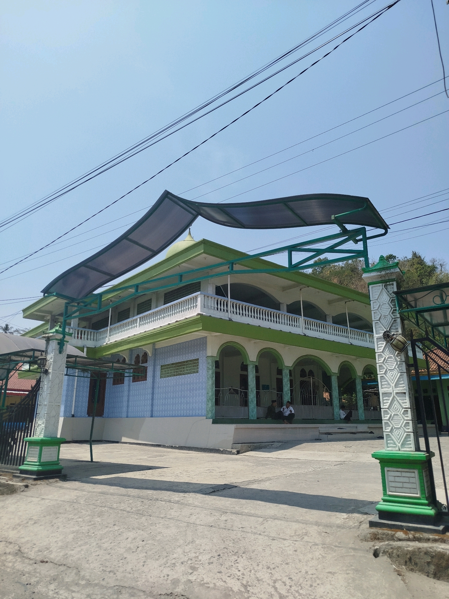 The image depicts the exterior of a multi-story building with a light blue and green facade, likely a mosque, in Kutosari, Indonesia. The building features arched openings on the ground floor, a balcony with a white railing on the second floor, and a green dome on the roof. A light gray corrugated roof structure is supported by green metal beams, extending over a portion of the building. Several people are seated on a raised platform in front of the building. The foreground shows a concrete ground surface with some debris and shadows. The background consists of a clear blue sky, overhead power lines, and a densely vegetated hillside. The scene is lit by natural daylight.