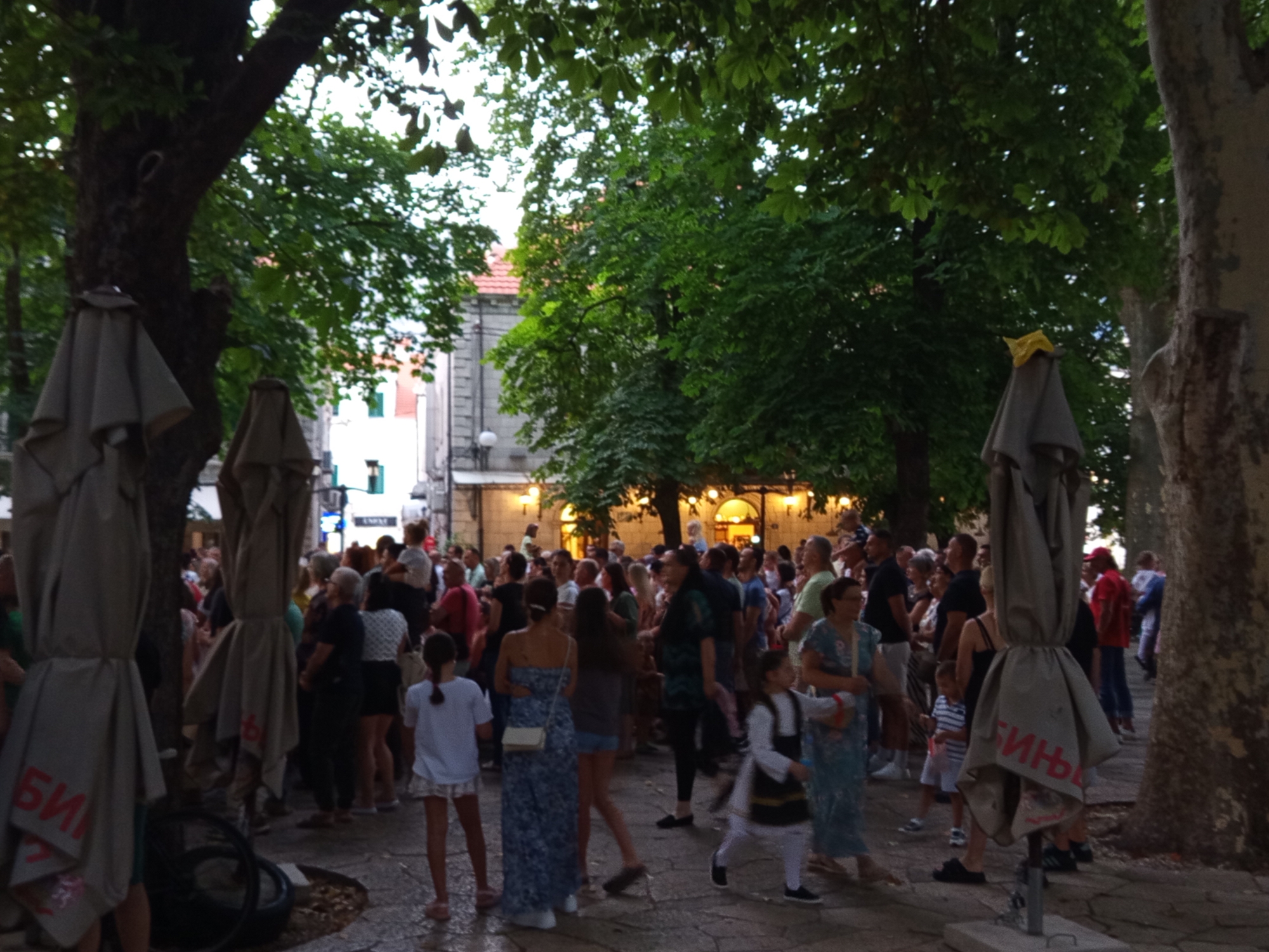 The image captures a lively outdoor gathering in Trebinje, Bosnia and Herzegovina, likely on a cobblestone street lined with trees and buildings. A crowd of people, varying in age and attire, are milling about, suggesting a festival or social event. Several large, closed umbrellas or canopies are prominent in the foreground, some with red lettering visible. In the background, a building with arched windows and illuminated lights indicates a commercial or public space, possibly a restaurant or cafe. The overall atmosphere appears relaxed and festive, with individuals engaged in conversation and observation. The presence of trees with lush green foliage suggests a warm season.