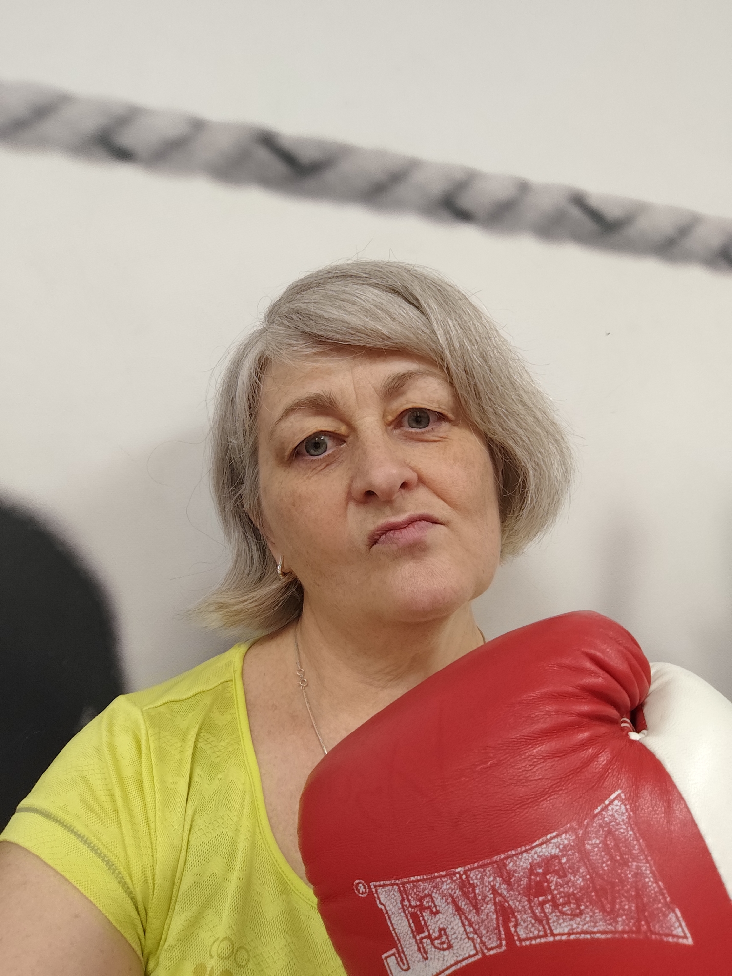 A middle-aged Caucasian woman with short, gray hair and fair skin is pictured in a medium shot. She is wearing a bright yellow t-shirt and a red and white boxing glove on her right hand, which is held up to her face. Her expression is serious, with her mouth slightly pursed and her eyes looking directly at the camera. The background appears to be a white wall with a rope-like design across the top. The lighting suggests an indoor setting, possibly a gym or sports facility. There is no information in the image to suggest a location in Krasnodar, Russia.