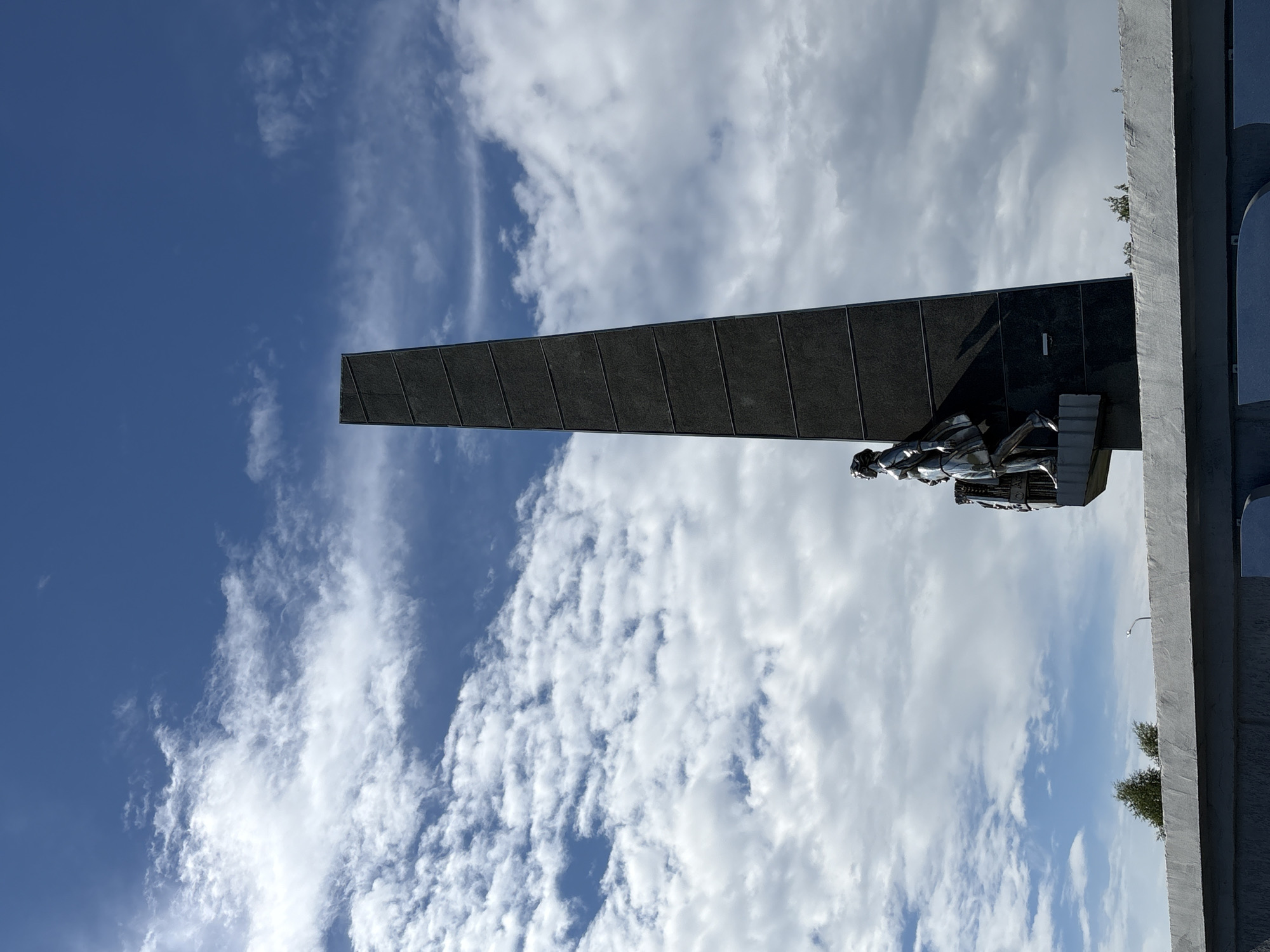 A monument with a tall, dark, obelisk-shaped structure stands against a bright blue sky with scattered white clouds. A silver sculpture of a woman walking is positioned at the base of the obelisk. The monument is situated on a concrete platform, with more concrete structures visible in the foreground. Trees are visible in the background. This is the monument "Per Aspera Ad Astra" in Kirovo-Chepetsk, Russia.