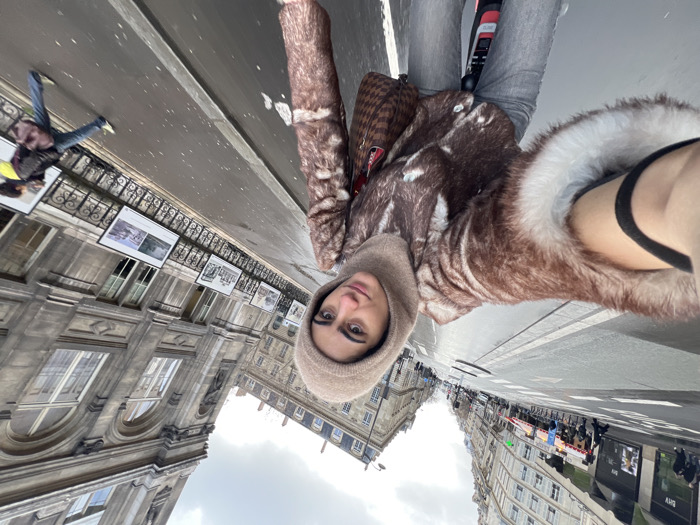 A young woman takes a selfie on a bicycle in Paris, France, near the BHV Marais department store, identifiable by the "BHV" sign on a building to the left. The scene is captured from a first-person perspective, with the woman centrally positioned, extending her right arm to hold the camera. She wears a brown and white patterned faux fur jacket with white cuffs, light grey jeans, and a light brown balaclava covering her head and neck. A brown checkered shoulder bag is visible on her left side.
The environment depicts a wet urban street under an overcast sky. To the left, a busy street with distinct lanes is bordered by multi-story Haussmannian-style buildings. To the right, a large, ornate gray stone building with tall arched windows features an outdoor exhibition of framed photographs displayed on a black metal railing along its base. A pedestrian, wearing blue jeans and a dark jacket and carrying a dark bag, walks away from the camera on the right side of the street, exhibiting motion blur. The foreground indicates the woman is on a bicycle, with a red frame element visible below her.