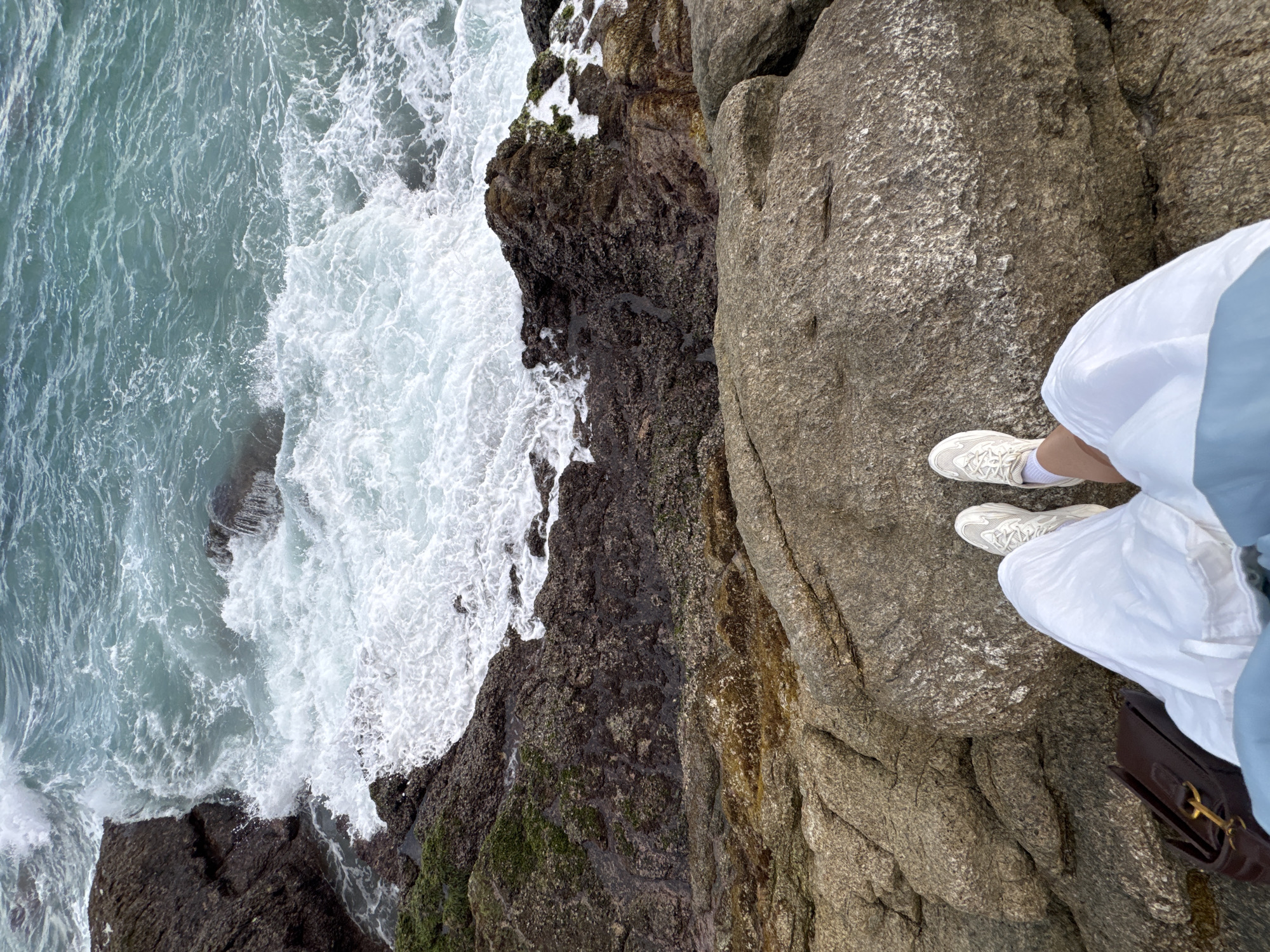 The image shows a high-angle view of a rocky coastline with waves crashing against the shore. In the foreground, a person's legs are visible, wearing white sneakers and a flowing white skirt. A dark brown handbag is placed on the rocks next to them. The sea is a vibrant turquoise, with white foam from the waves. The rocks are varied in color and texture, with some appearing dark and wet, covered in moss, while others are lighter and rougher. The scene appears to be a natural coastal environment, possibly near Choeng Thale, Thailand, given the tropical-looking water and vegetation on the rocks.