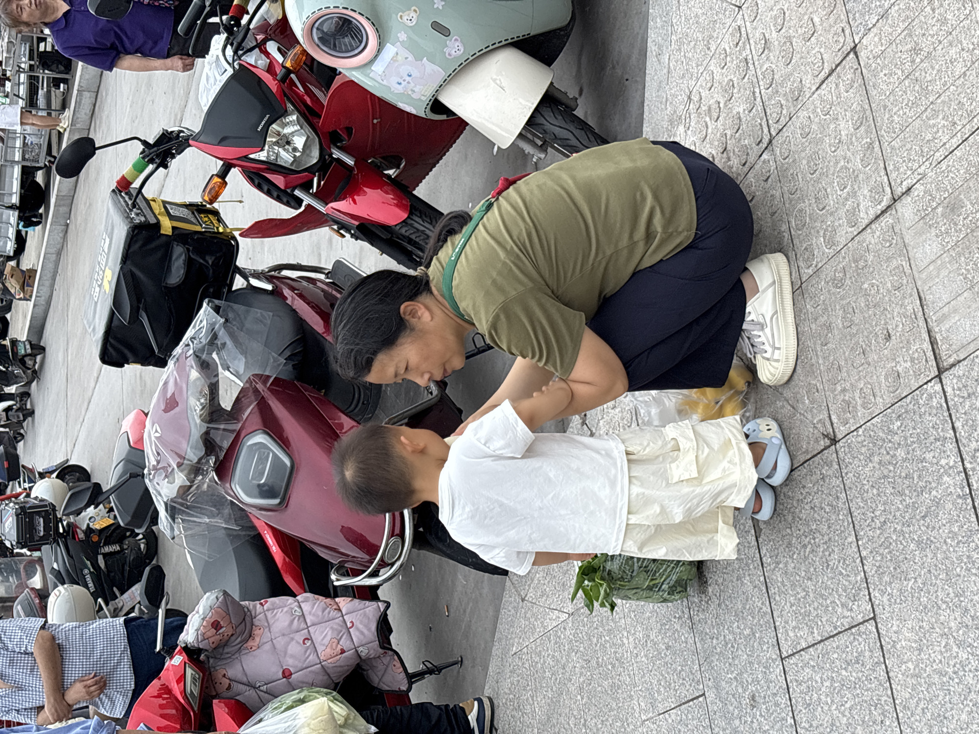 A woman kneels on a paved surface, interacting with a young child dressed in white. The woman is wearing a green shirt, dark pants, and white shoes. Multiple scooters and motorcycles are parked nearby, partially obscured by covers and bags. The scene appears to be outdoors, on a city street. The presence of scooters and packaging suggests a commercial or residential area. It is in the city of Jiangjin, China.