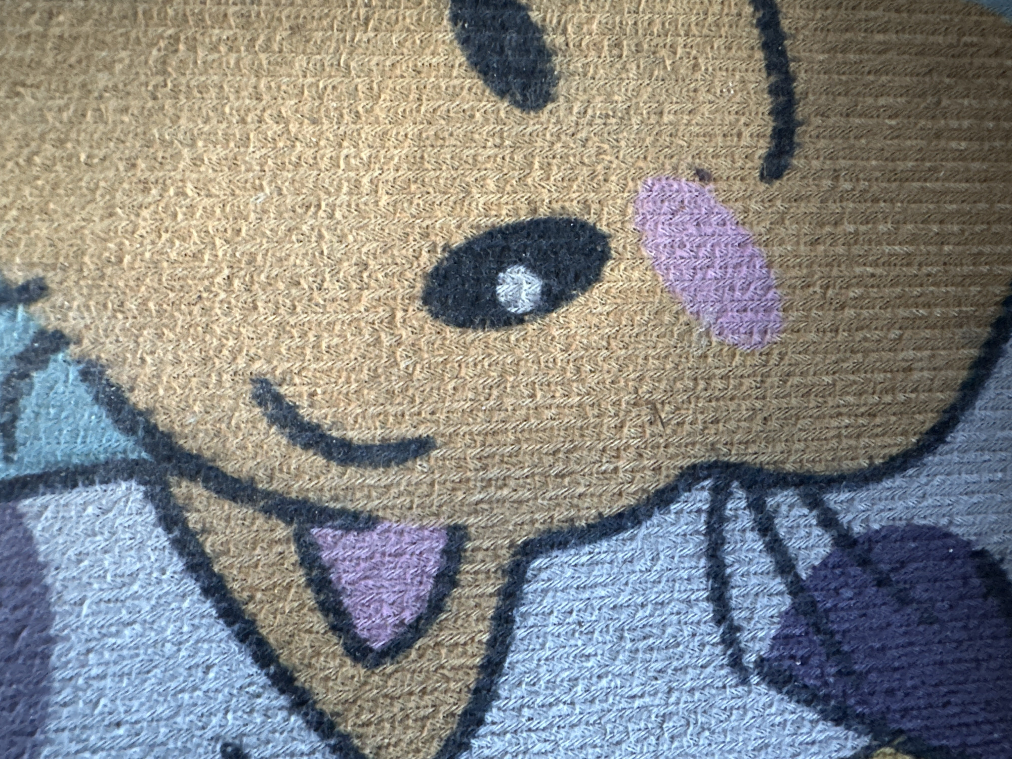 The image shows a close-up of a woven fabric, likely a rug or blanket, featuring a cartoon character. The character has a brown face with a black eye containing a white highlight, and a light pink cheek blush. One ear is visible, outlined in black and filled with light purple. Part of the character's body is in a light blue and purple hue, also outlined in black. The background fabric has a visible woven texture with a pattern of small raised loops. There are no discernible human subjects or actions. The location is not specified in the image.