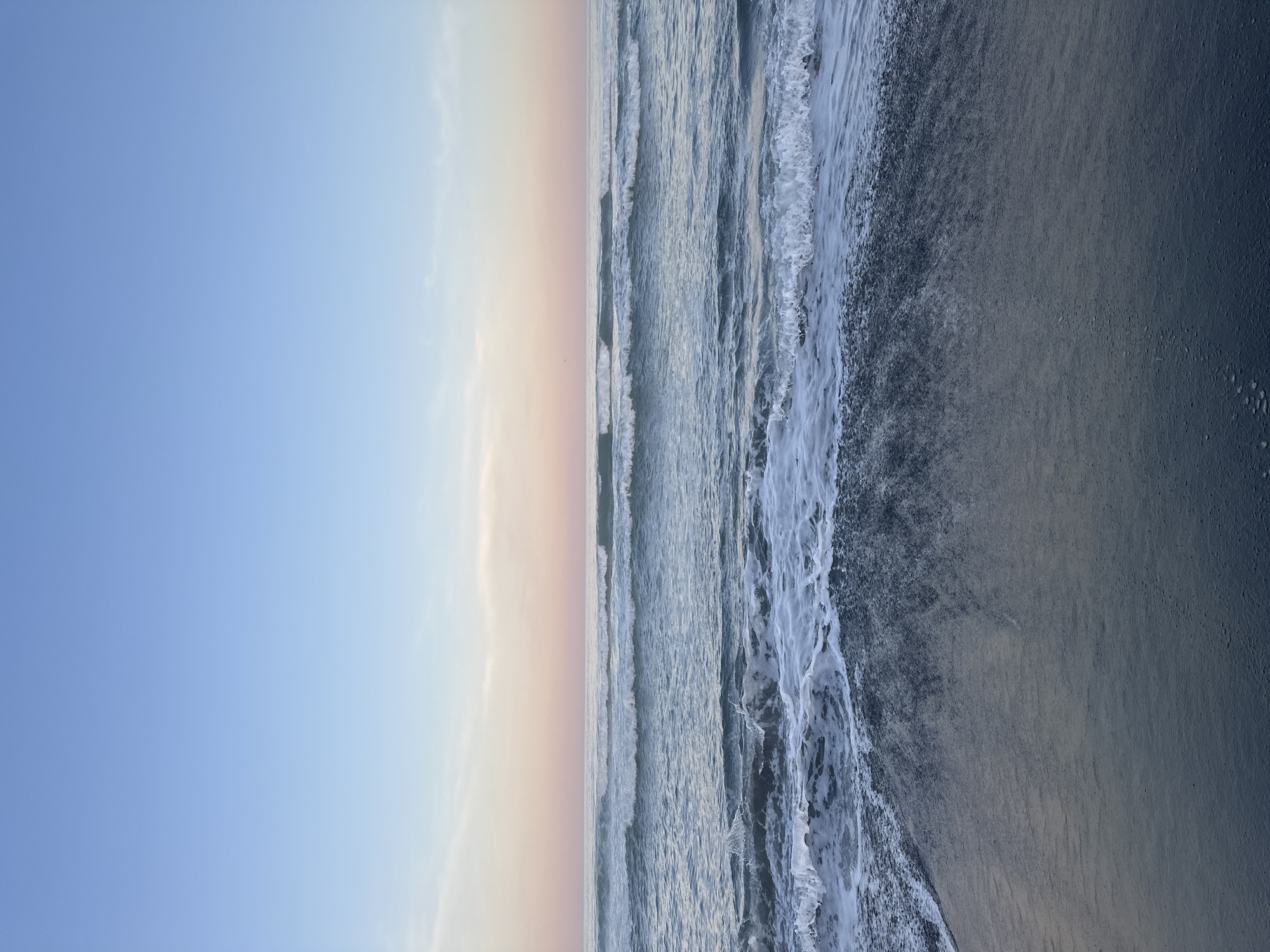 The image depicts a tranquil ocean scene, likely at dusk or dawn, with a clear sky transitioning from a pale blue at the top to soft pink and peach hues near the horizon. Gentle waves roll towards a dark, wet sand beach. The water is a deep blue-green, with white foam cresting on the waves closest to the shore. There are no people or man-made objects visible in the image. The environment is natural and serene. This scene is consistent with the coastal areas surrounding Petropavlovsk-Kamchatskiy, Russia, which is known for its dramatic volcanic landscapes and Pacific Ocean coastline.