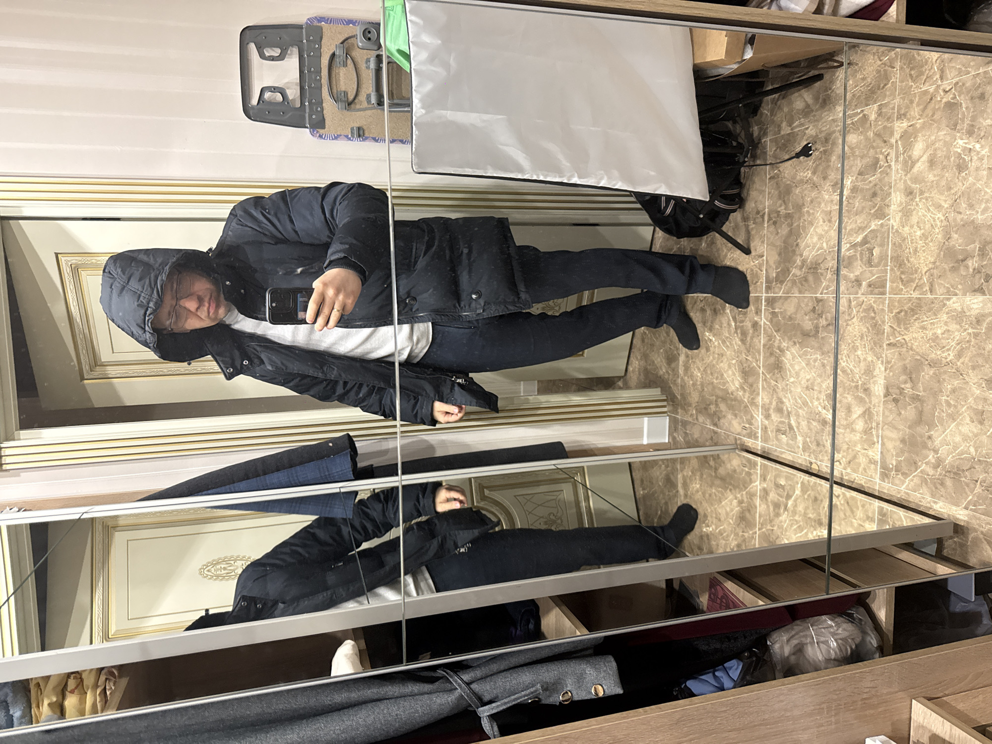 A man in Khimki, Russia, takes a selfie in a mirrored wardrobe. He wears a hooded dark blue winter coat, a white shirt, dark blue trousers, and dark socks. The wardrobe doors are mirrored, reflecting his image. Behind him, on the wardrobe's wall, is an ironing board and a white reflector. The wardrobe's interior contains clothing items in drawers. The background features a marble-patterned wall behind the wardrobe.